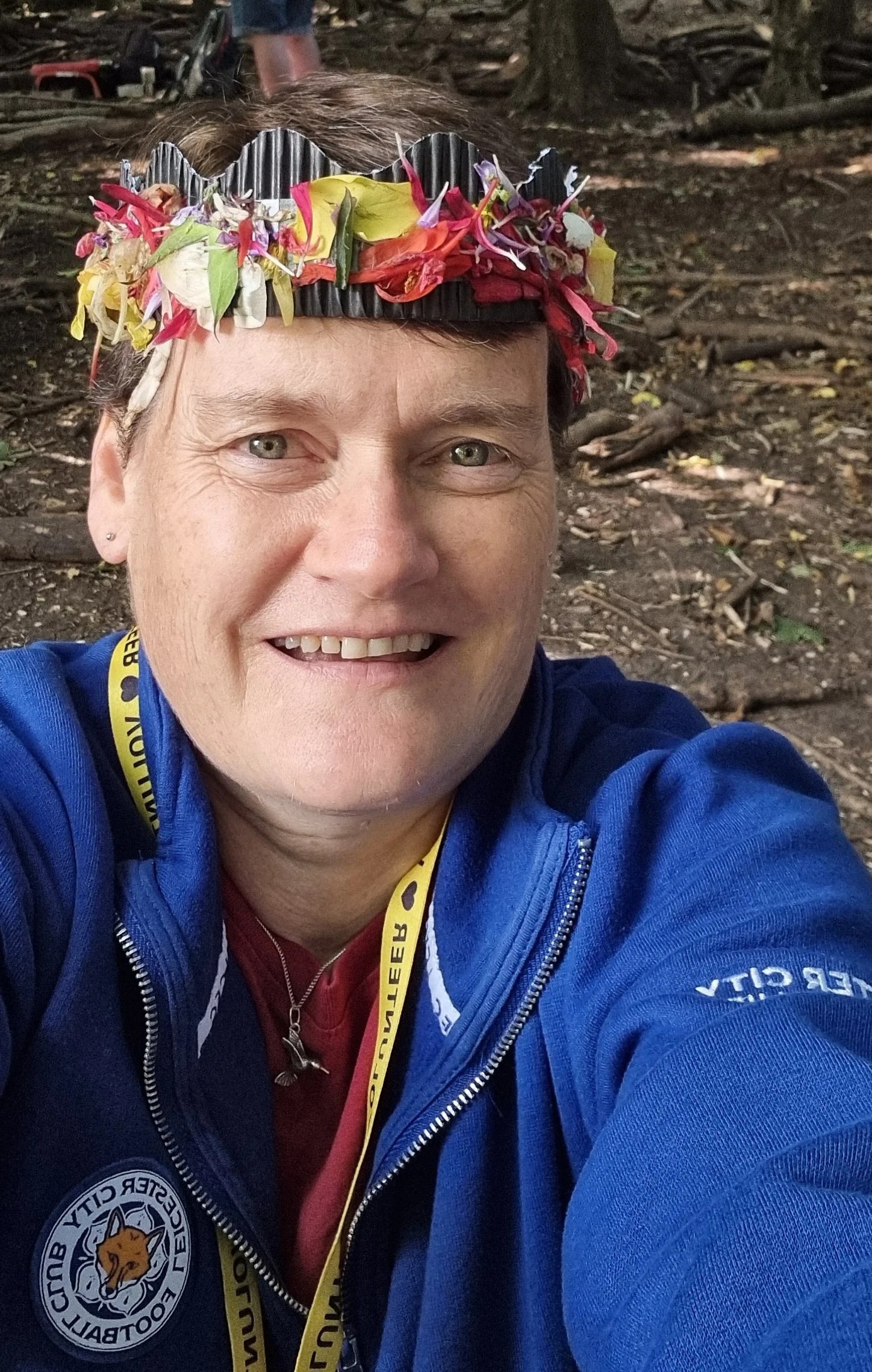 A person wearing a blue jacket with a lion logo, a yellow lanyard, and a colorful flower crown, smiling outdoors in a wooded area.