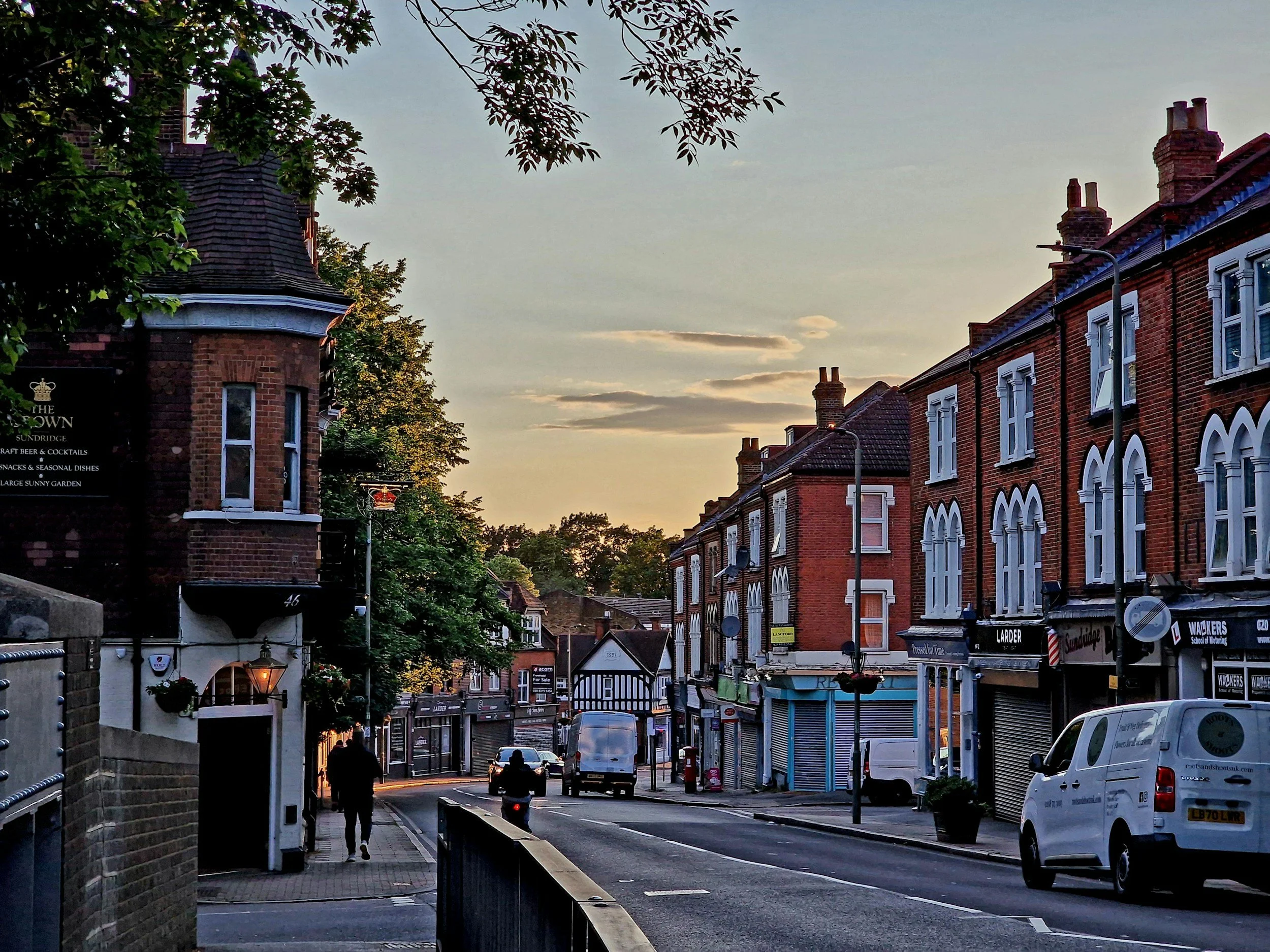 A street scene at sunset in a small town with brick buildings, parked vehicles, and a pedestrian walking down the sidewalk.
