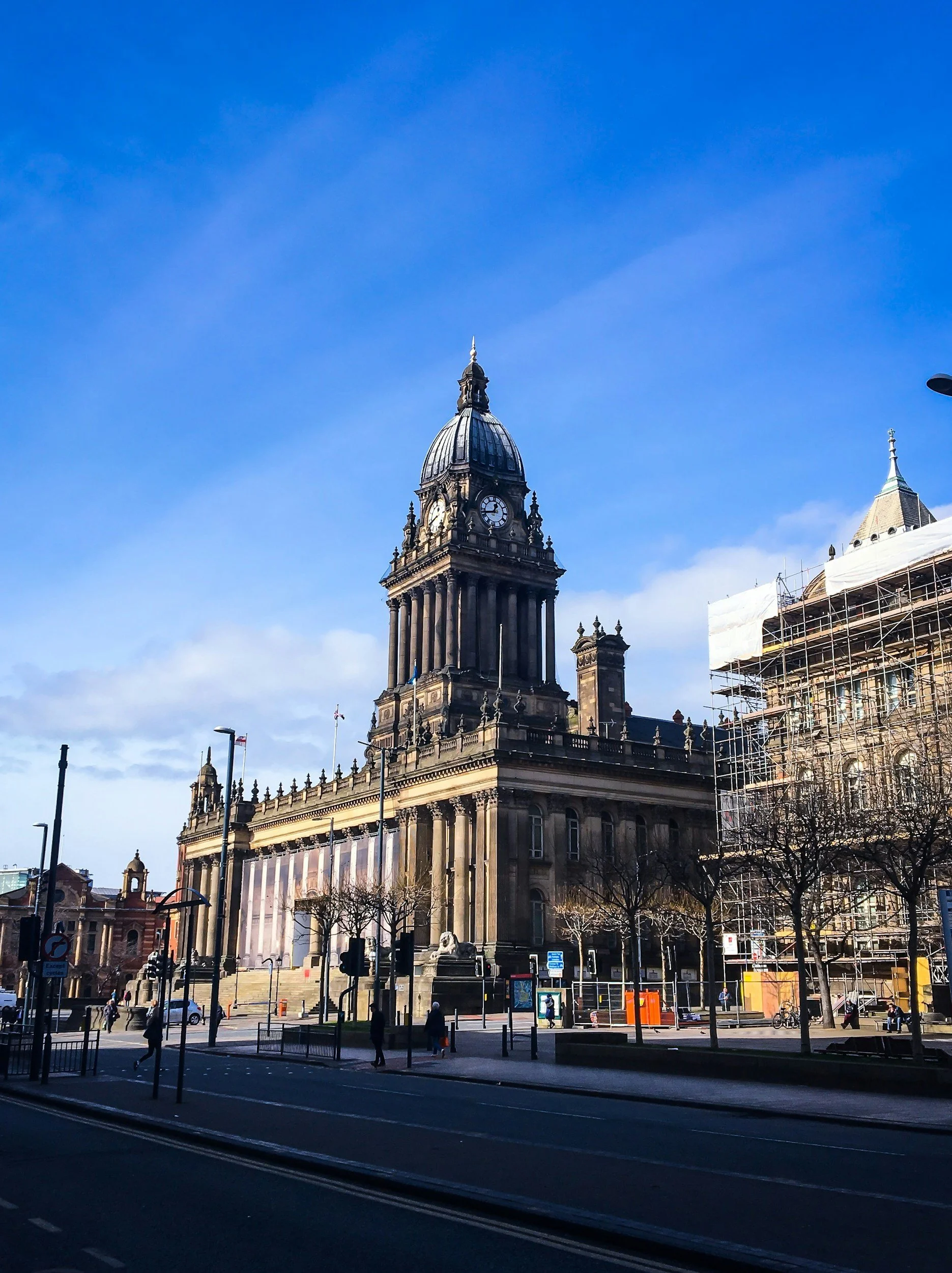 A historic city hall building with a clock tower, featuring a domed roof, surrounded by leafless trees and city streets during daytime with clear blue sky.