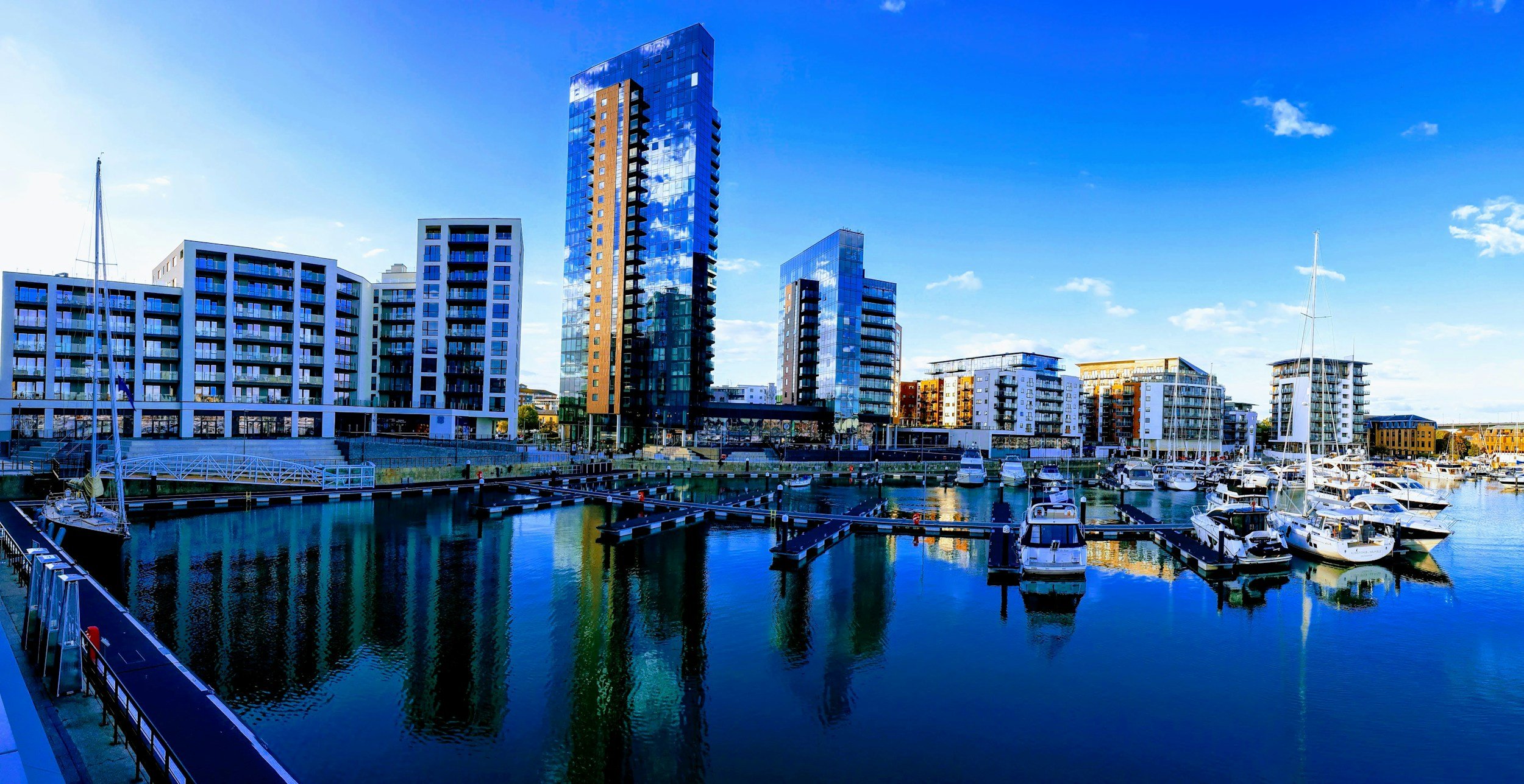 A modern cityscape with tall glass buildings, a marina filled with sailboats, and a clear blue sky reflection on the water.
