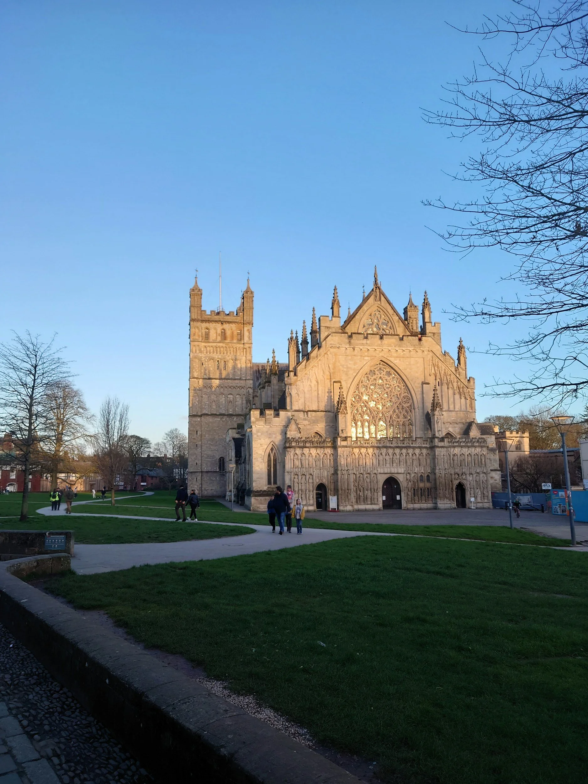 Exterior view of a historic Gothic cathedral with a large rose window, pointed arches, and ornate stone carvings, set against a clear blue sky. People are walking on the surrounding pathways and grassy areas.