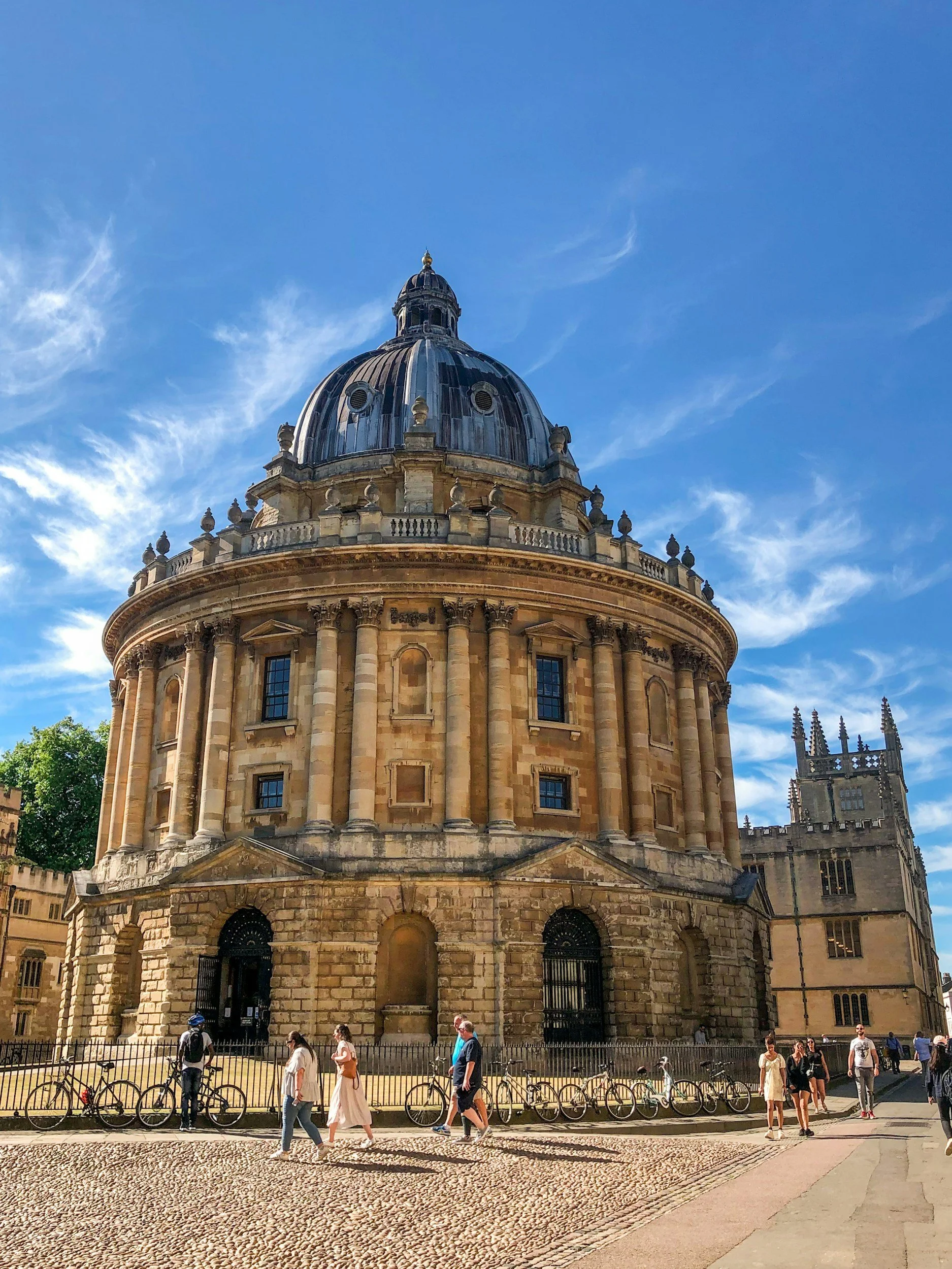 The historical building with a large dome and classical architecture, pedestrians walking nearby, bicycles parked along the fence, blue sky with clouds.