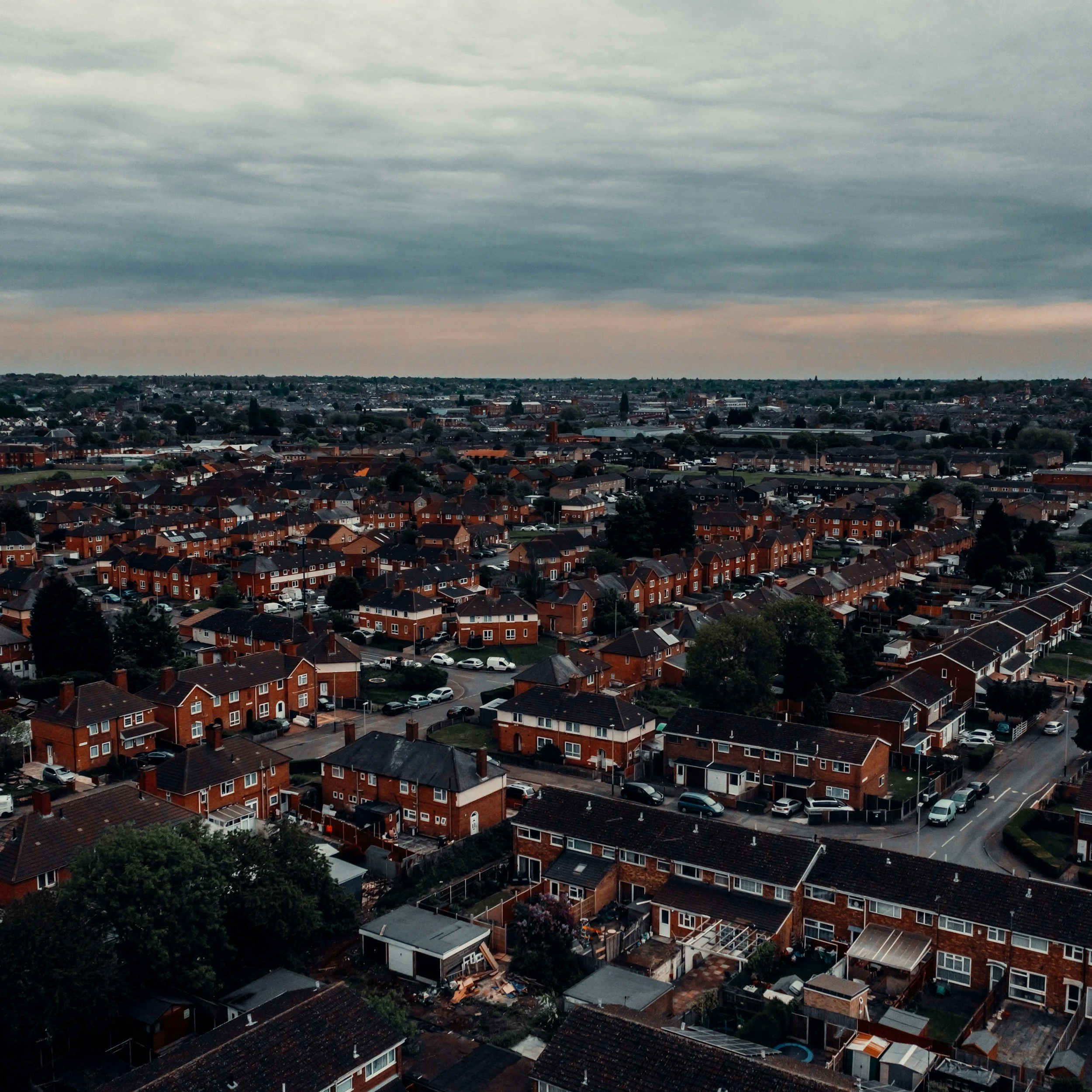 Aerial view of a suburban neighborhood with rows of brick houses, cars parked on the streets, and trees, under a cloudy sky during late afternoon or early evening.