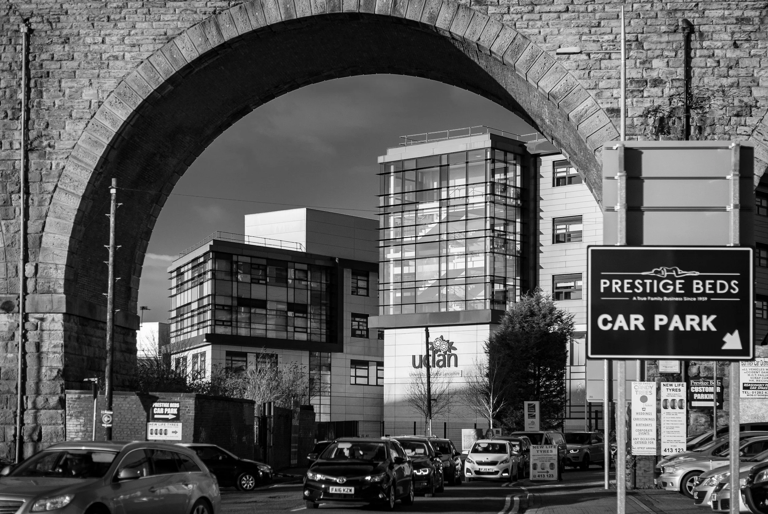 City street scene with cars parked and driving, a sign indicating car park for Prestige Beds, buildings in the background, and an old stone archway in the foreground.