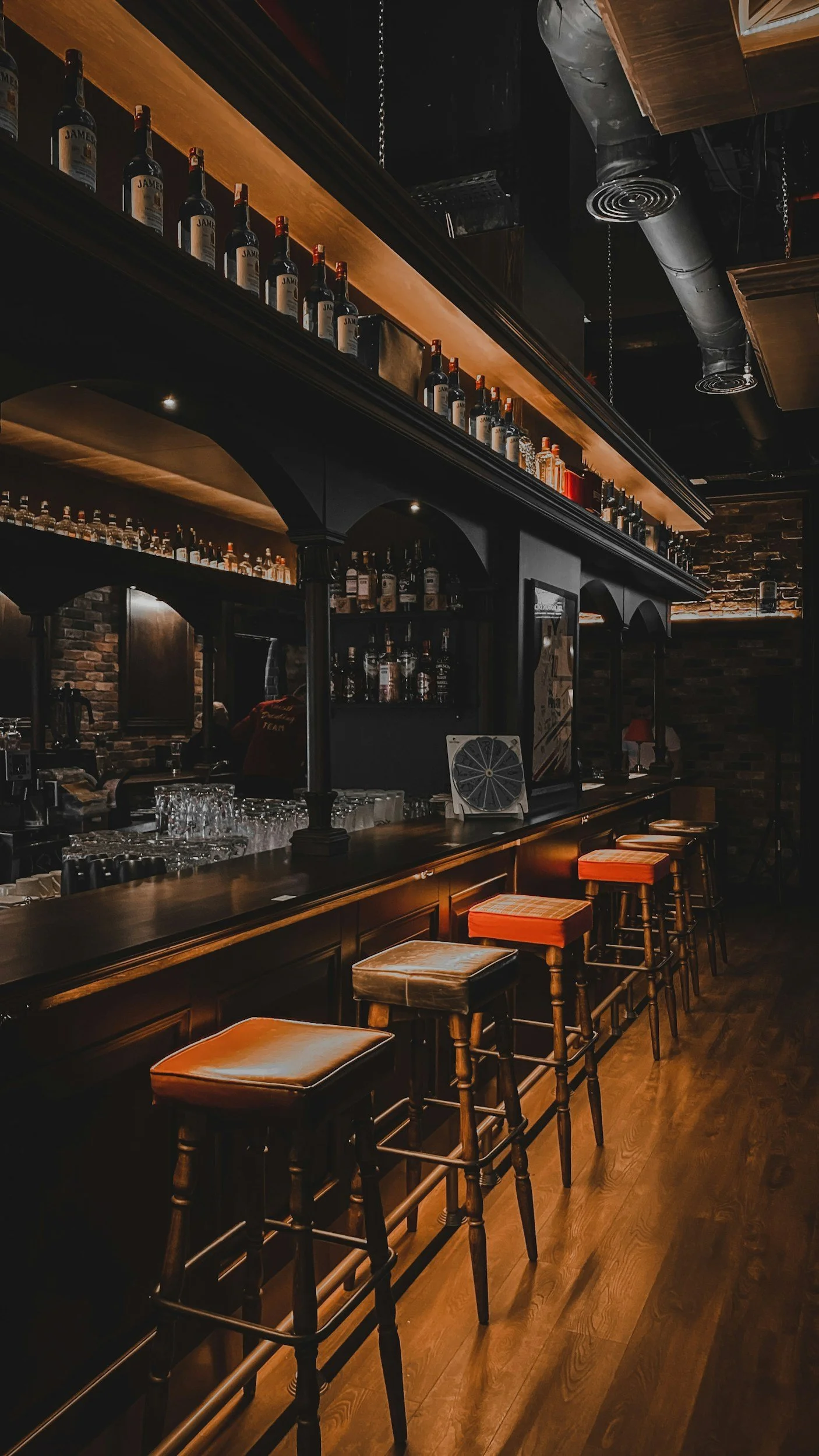 A dimly lit bar with a wooden counter and barstools with leather seats, dark wood accents, bottles of alcohol on shelves, glasses on the counter, and brick walls in the background.