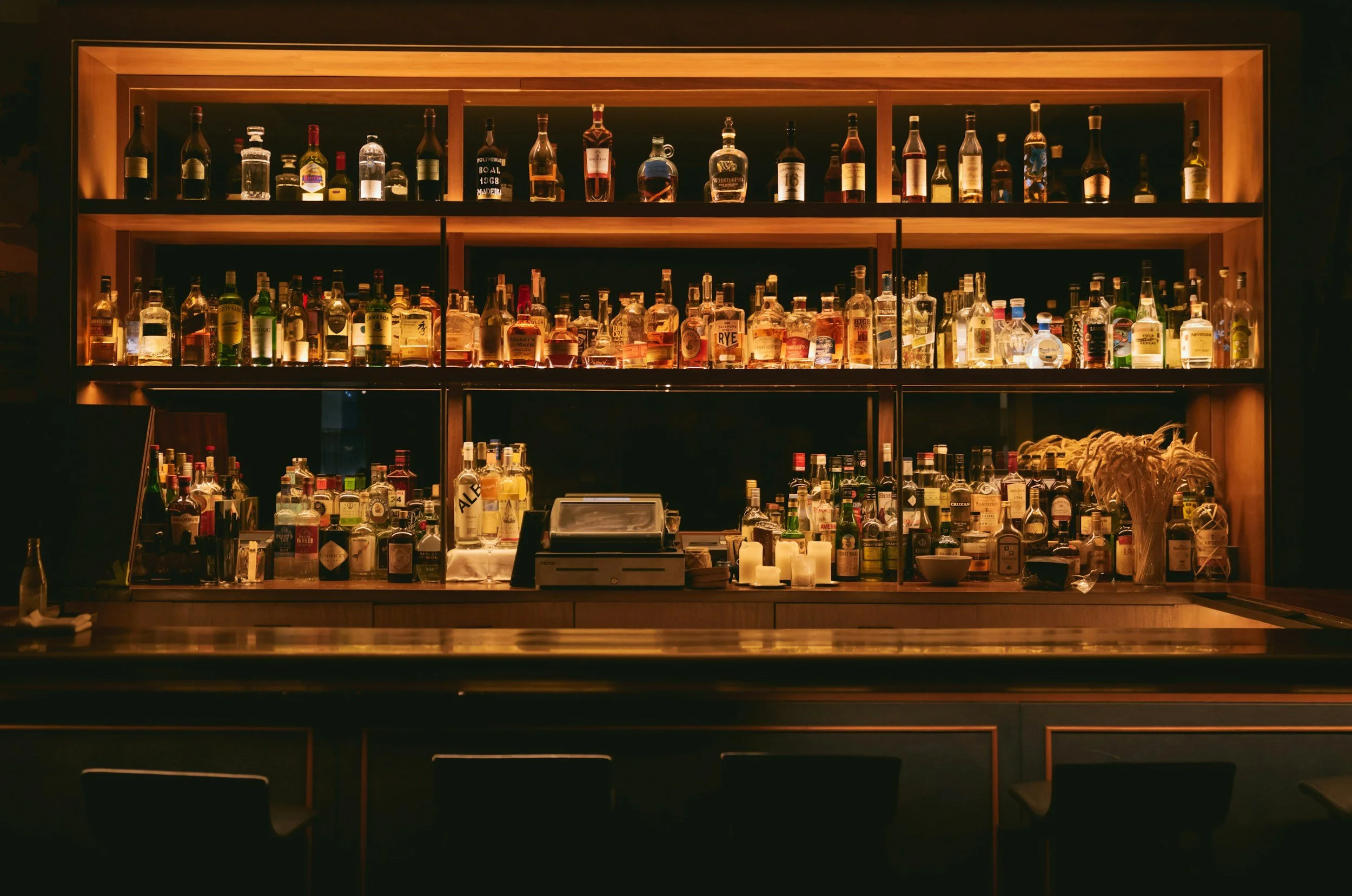 A well-lit bar with shelves of various liquor bottles on backlit wooden shelves behind the bar counter.