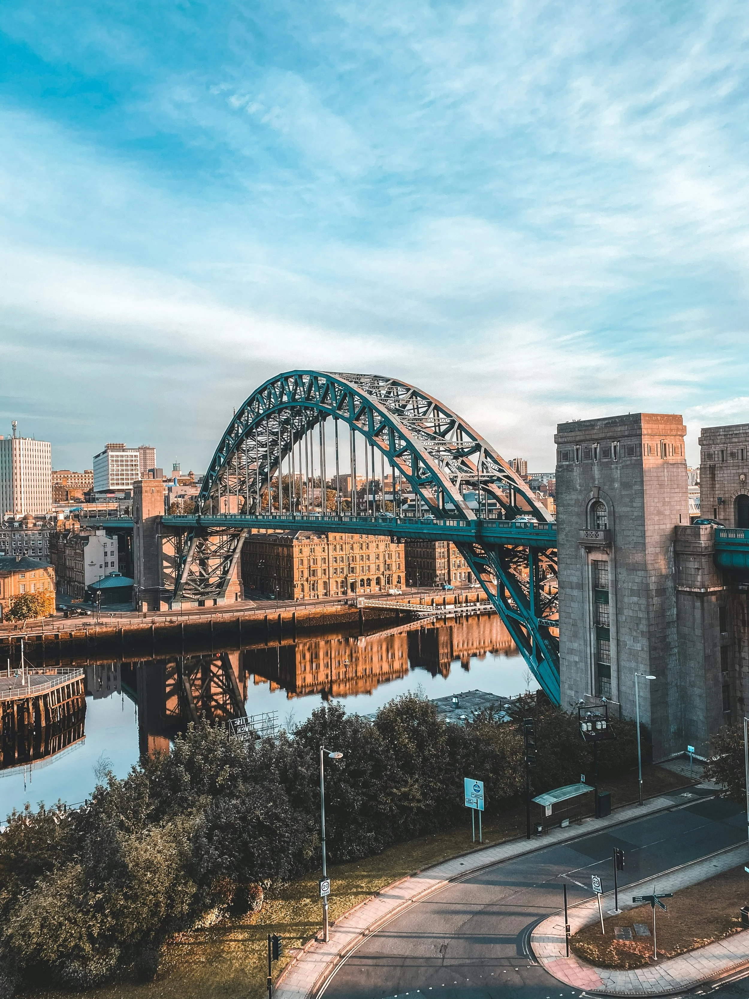 View of the Tyne Bridge in Newcastle, Australia, with the river reflecting the bridge and city buildings under a partly cloudy sky.