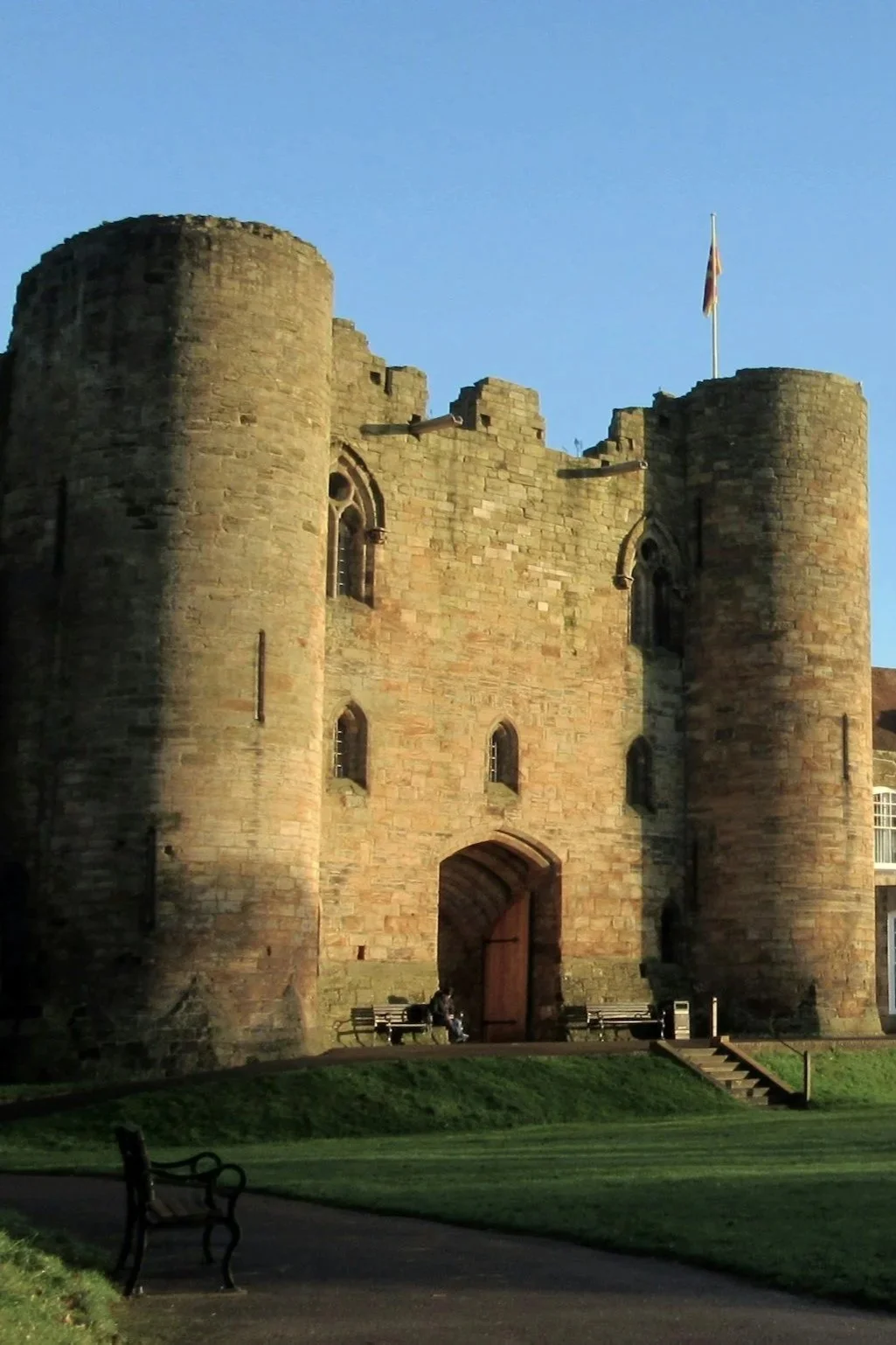 A historic stone castle with rounded towers and a wooden gate, featuring a British flag on top, surrounded by a park with benches and a grassy area.