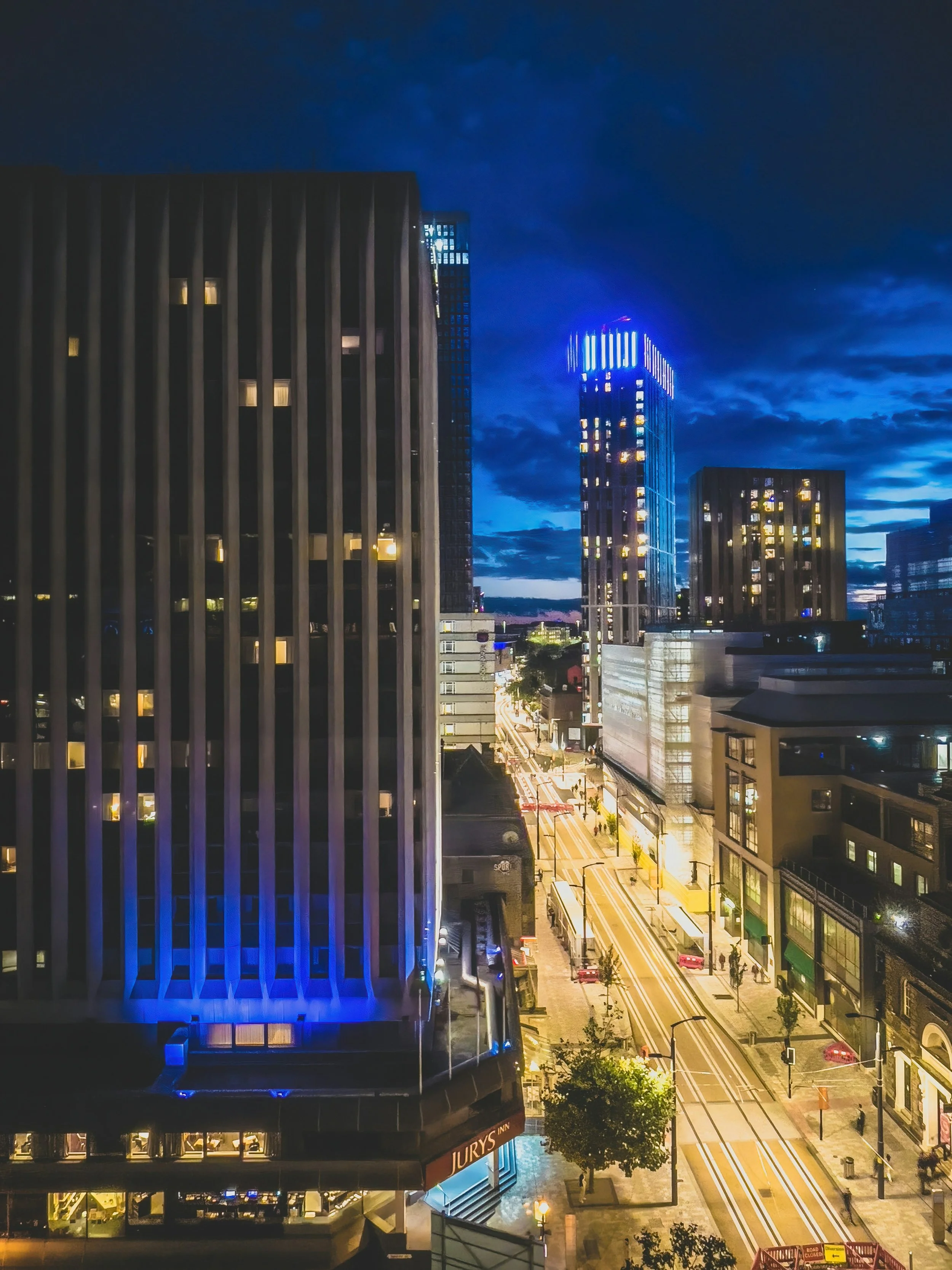 Nighttime cityscape view of tall illuminated buildings with streets below, some cars and streetlights, beneath a dark blue sky with clouds.