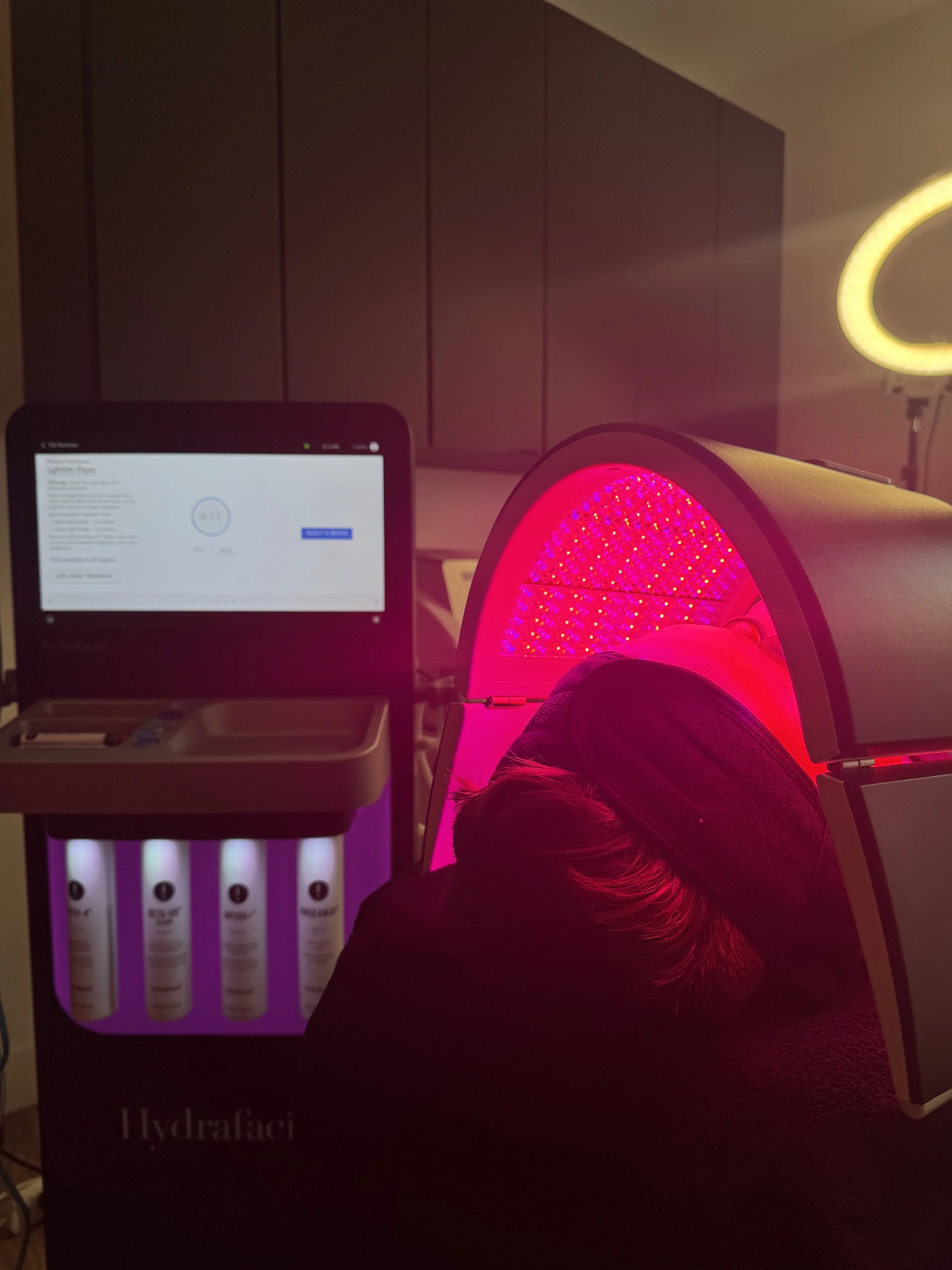 Person lying inside a red LED light therapy device, with a computer screen and product display in the background, labeled Hydrafacial.