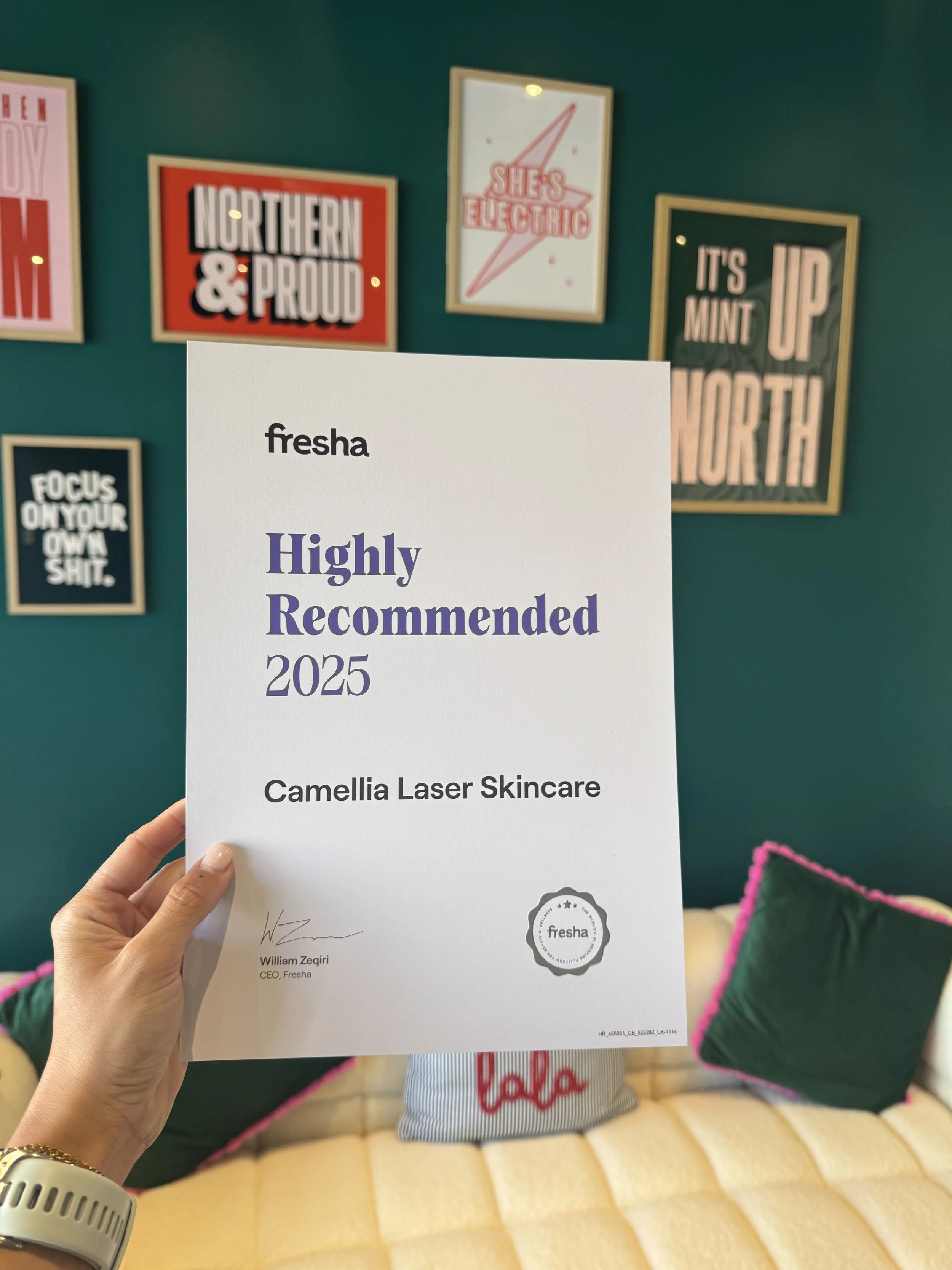A person holding a certificate from Fresha that reads 'Highly Recommended 2025 Camellia Laser Skincare' in front of a teal wall with framed motivational and pride-themed posters. The posters include phrases like 'Northern & Proud', 'She's Electric', 