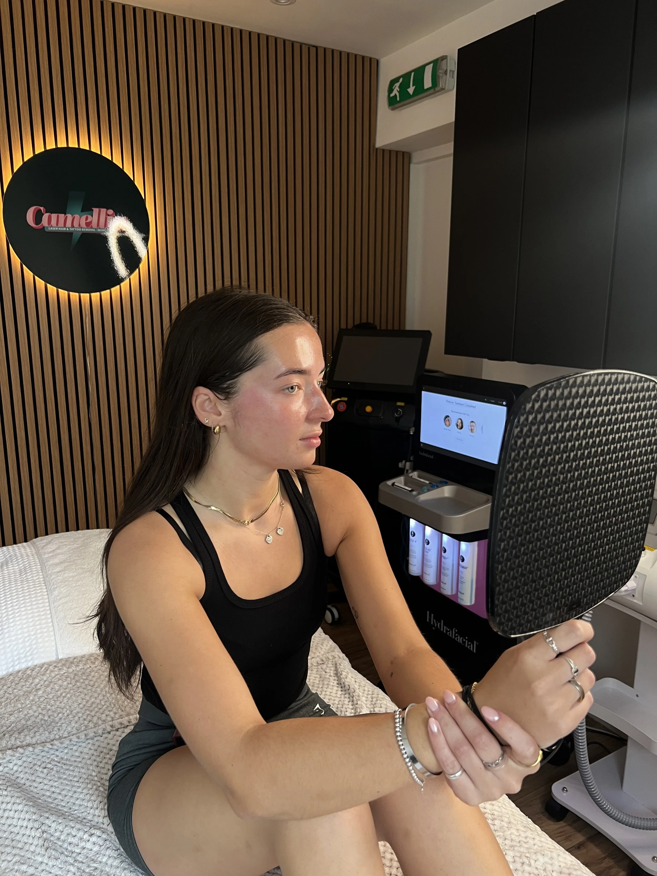 Young woman in black tank top and shorts sitting on a bed at a skincare clinic, holding a handheld device used for skin treatment.