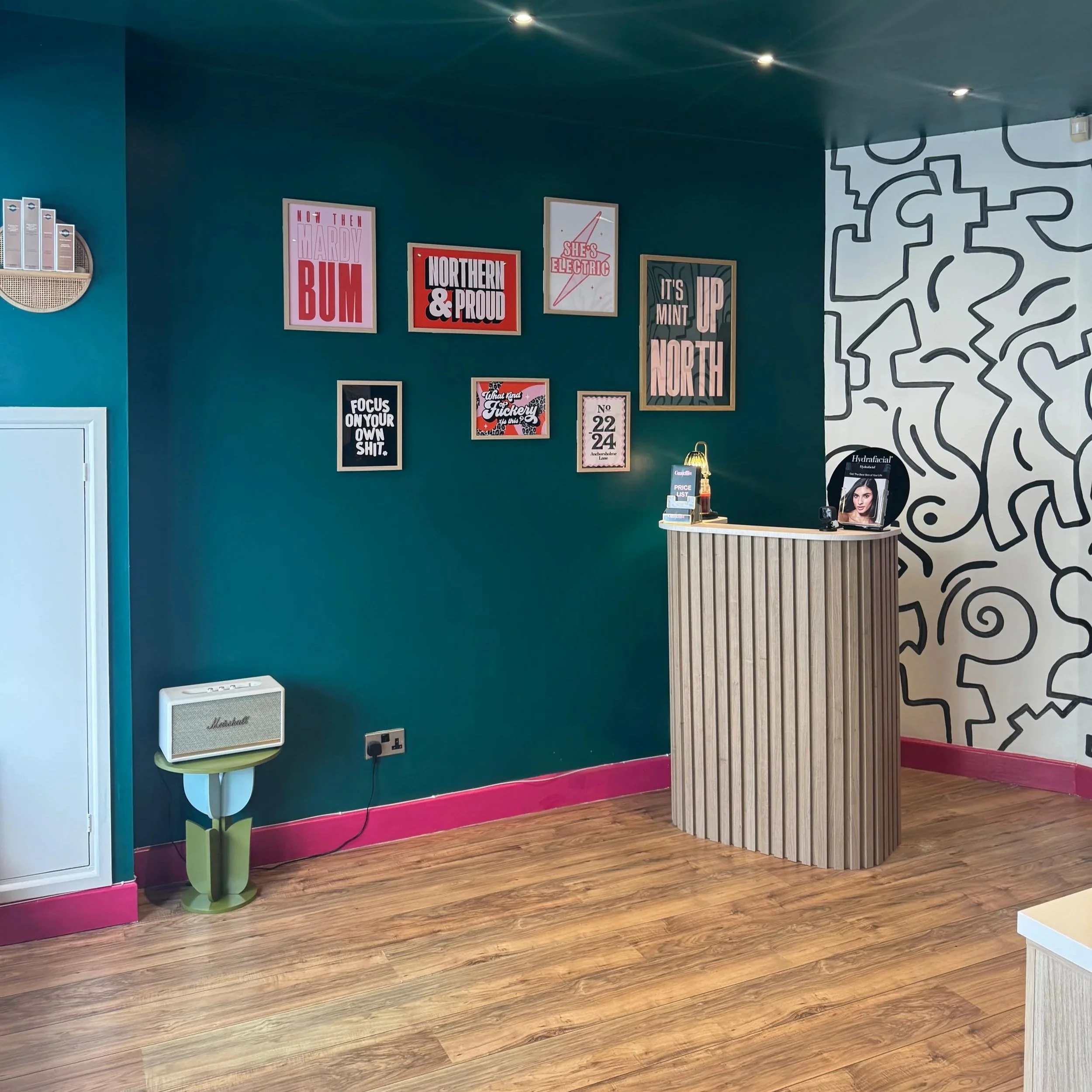 Interior of a room with a teal wall decorated with multiple framed posters and quotes, a small reception desk made of vertical wooden slats, a Marshall speaker on a modern green stand, and a black-and-white abstract mural on the right wall. The floor is wood, and there is pink trim along the base of the walls.