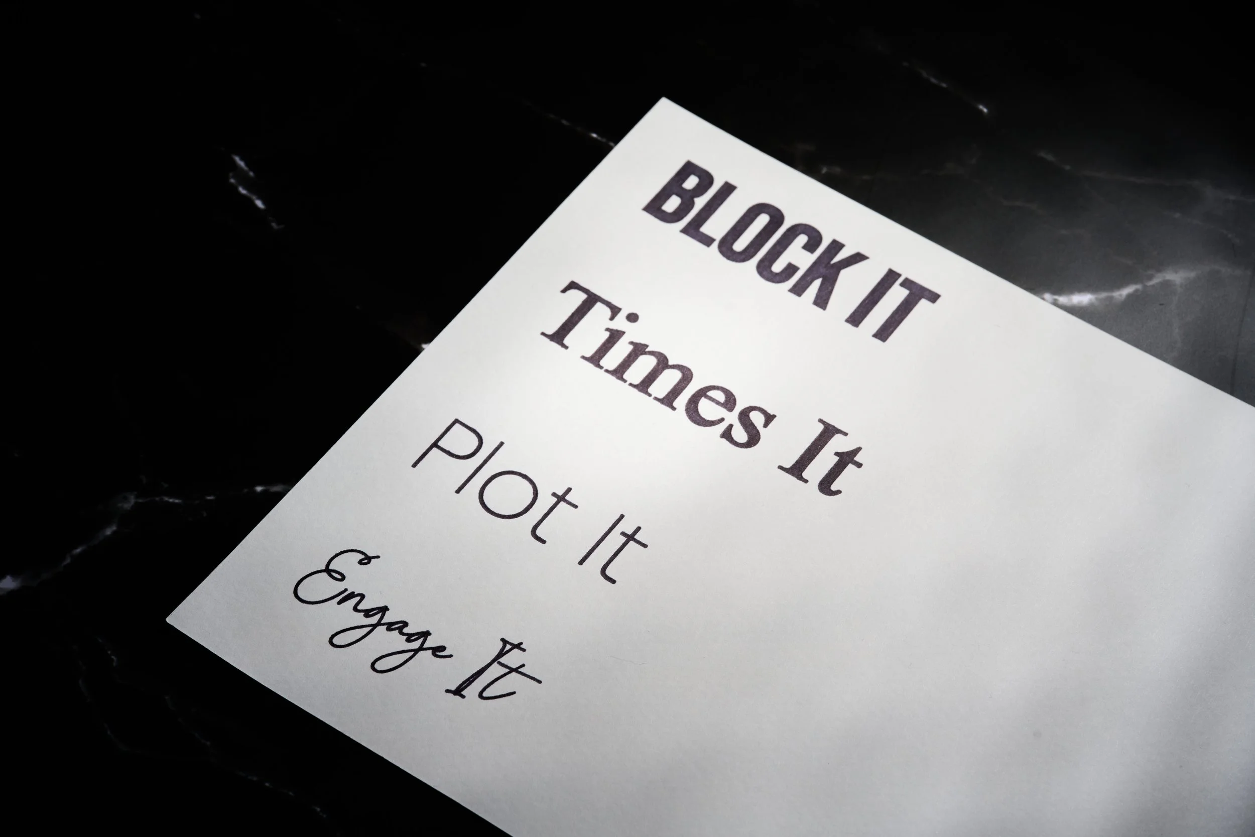 A printed sign says 'BLOCK IT,' 'Times It,' 'PLOT It,' and 'Engage It' on a white sheet of cold pressed watercolour paper placed on a dark marble surface.