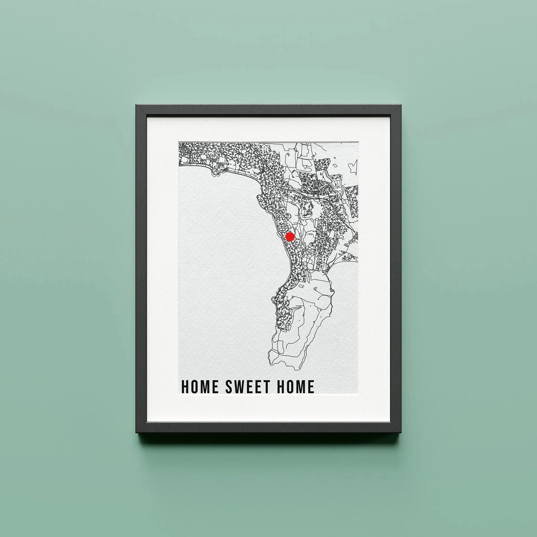 A framed black and white map of a city with a red dot marking a specific location, on a light green wall. Text below the map reads 'HOME SWEET HOME'.