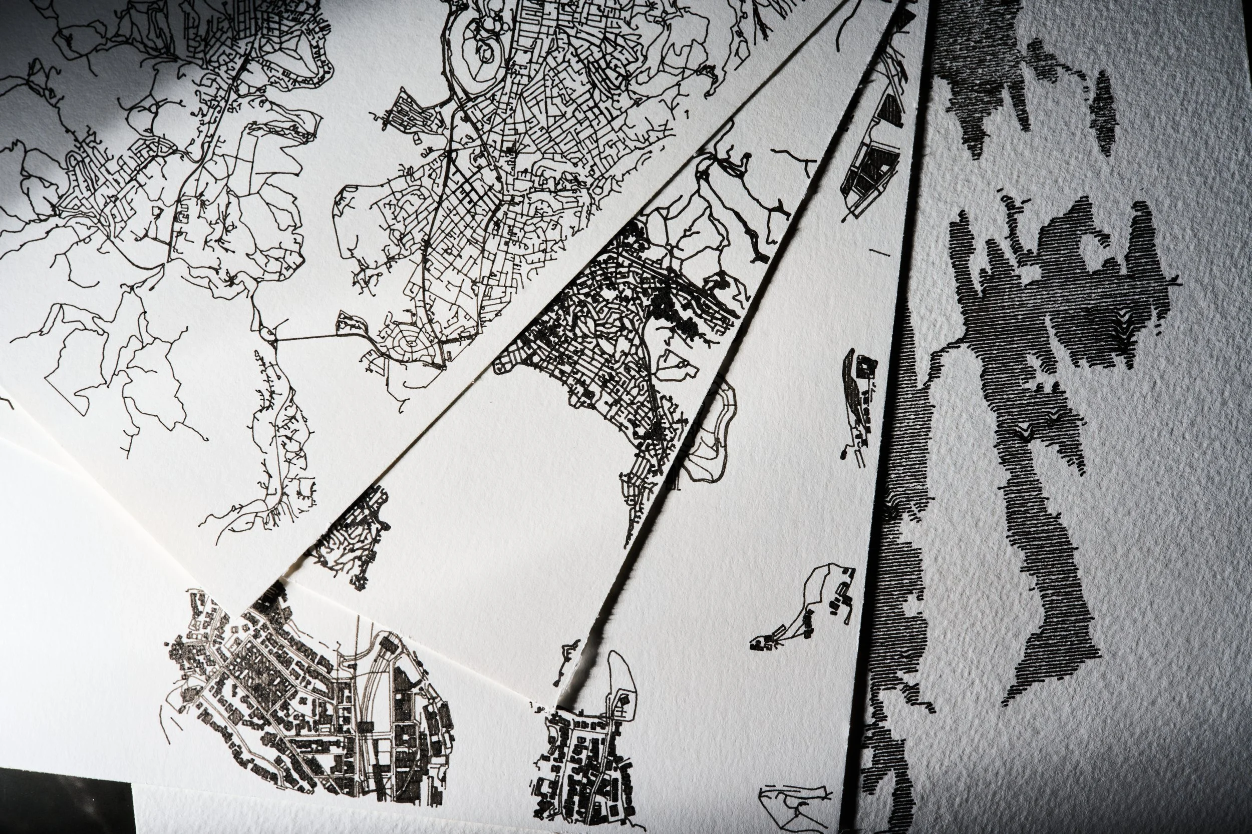 A collection of maps and illustrations drawn with black ink on textured white watercolour paper of different city layouts and geographic features layered together on a flat surface.