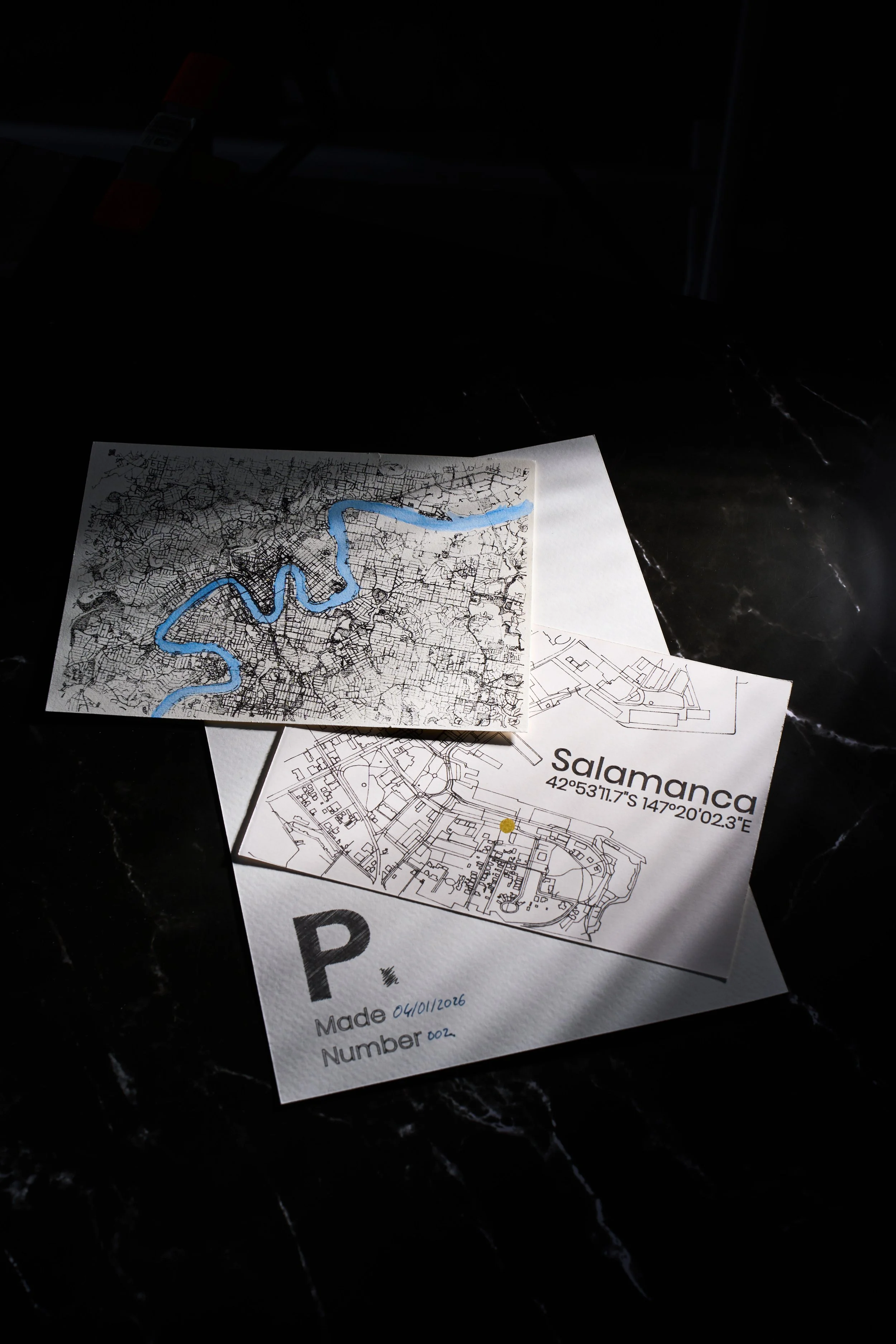 Maps and notes on a dark surface showing a city map labeled Salamanca with coordinates, an outline map with a highlighted river, and handwritten notes including a parking permit and date.