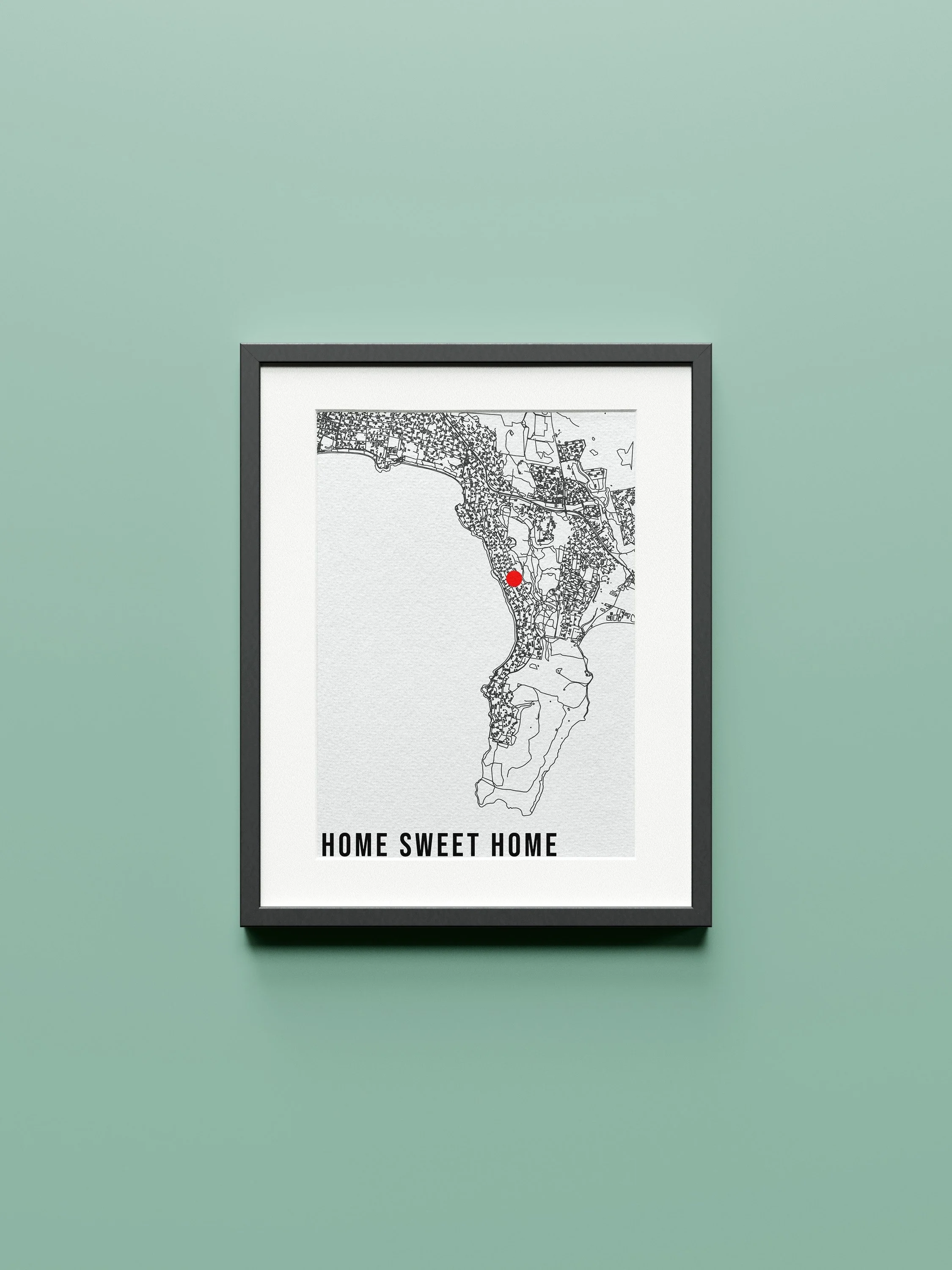 A framed map of a coastal city with a red dot marking a location, and the text 'Home Sweet Home' at the bottom, hanging on a pale green wall.