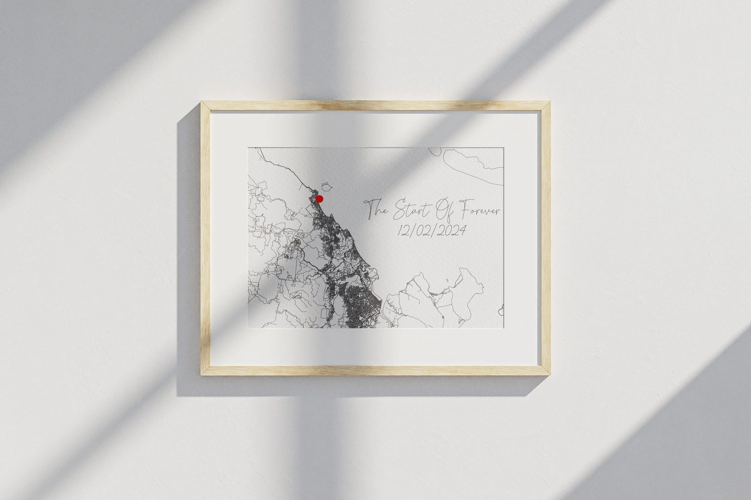 A framed map with a red dot on Thomas'  rough location in a city on the west coast of the United States. The frame hangs on a white wall with shadows cast across it. Handwritten text on the map reads 'The Start Of Forever 12/02/2024'.