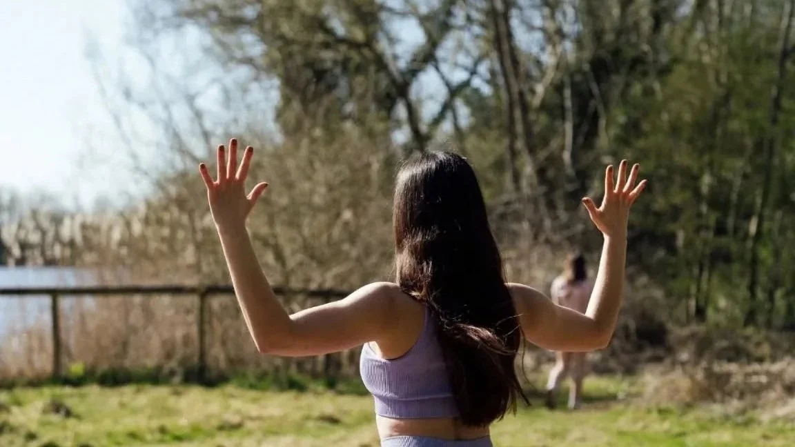 A woman with long dark hair wearing a purple crop top standing outdoors with her arms raised, back to the camera, in a natural setting with trees and another person in the background.