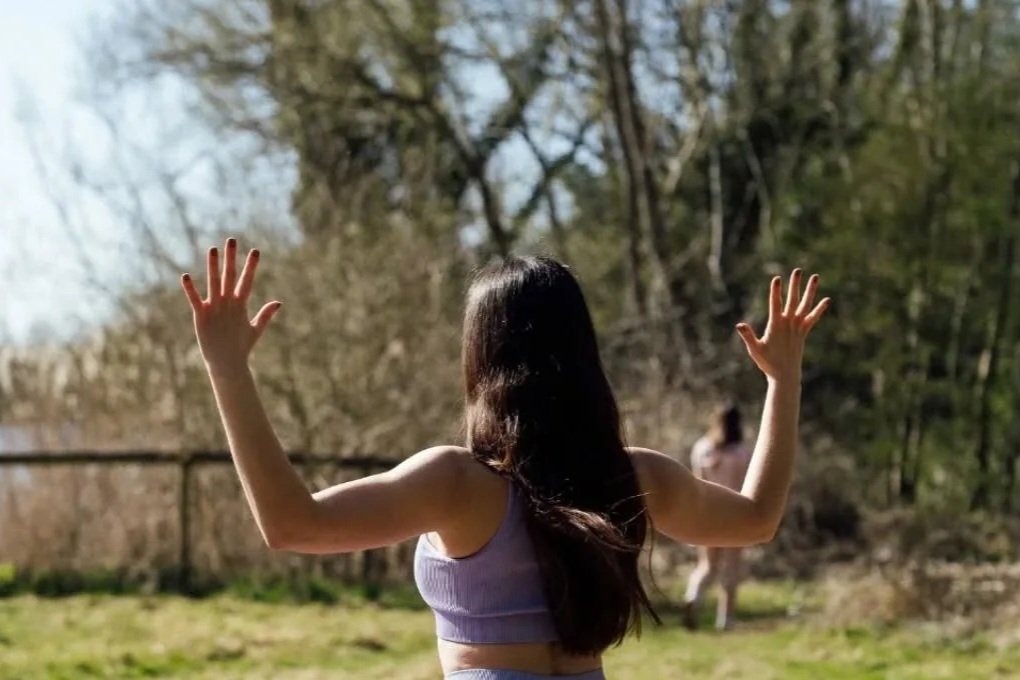 A woman with long dark hair facing away, arms raised outdoors during the daytime, with trees and a person in the background.