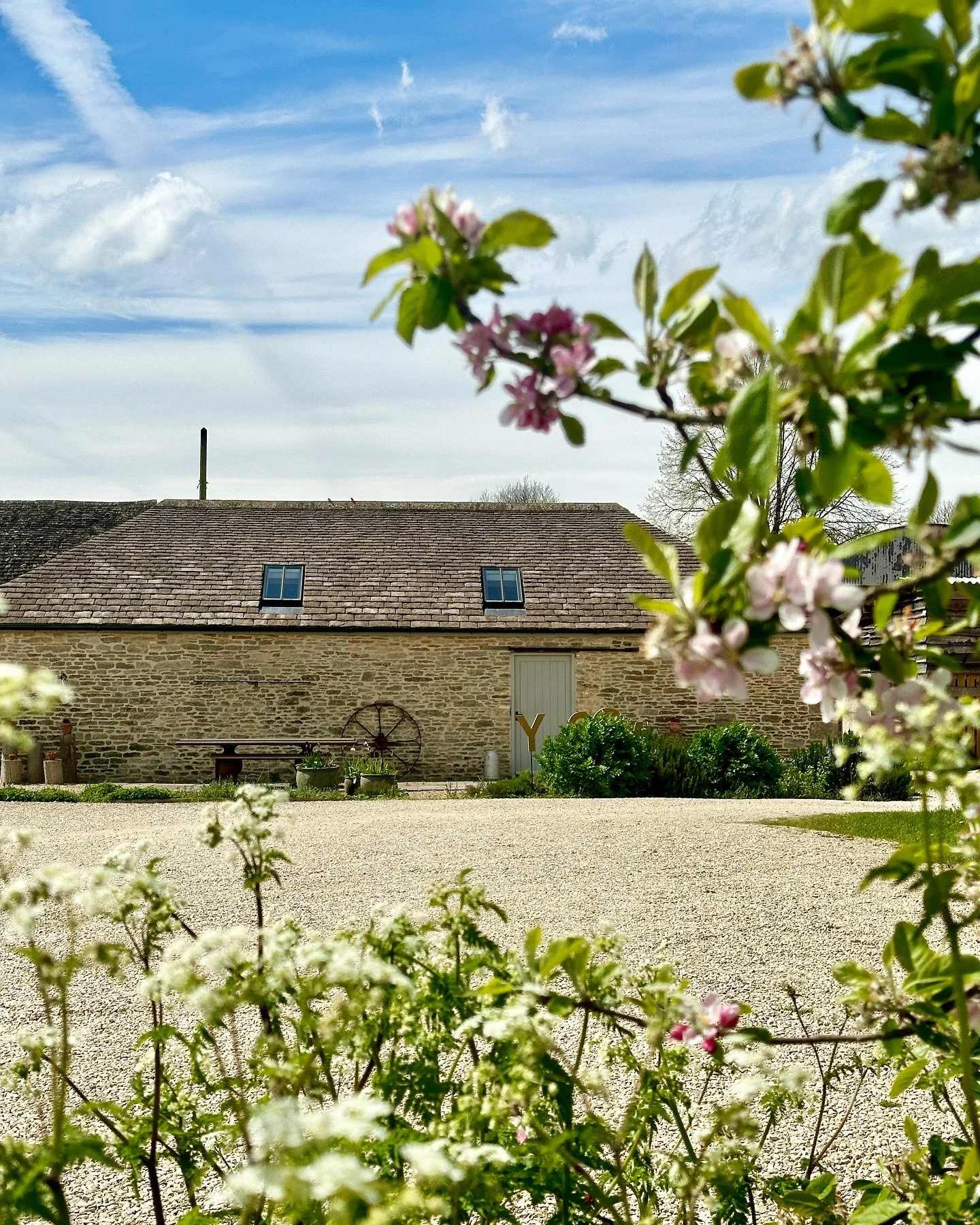 A stone house with a pitched roof and small windows, with pink and white flowers in the foreground, under a partly cloudy sky.