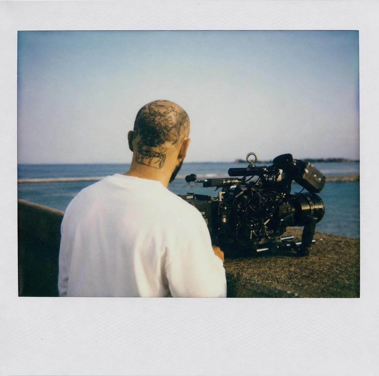 A man with a tattooed head operating a professional camera near the ocean during sunset.