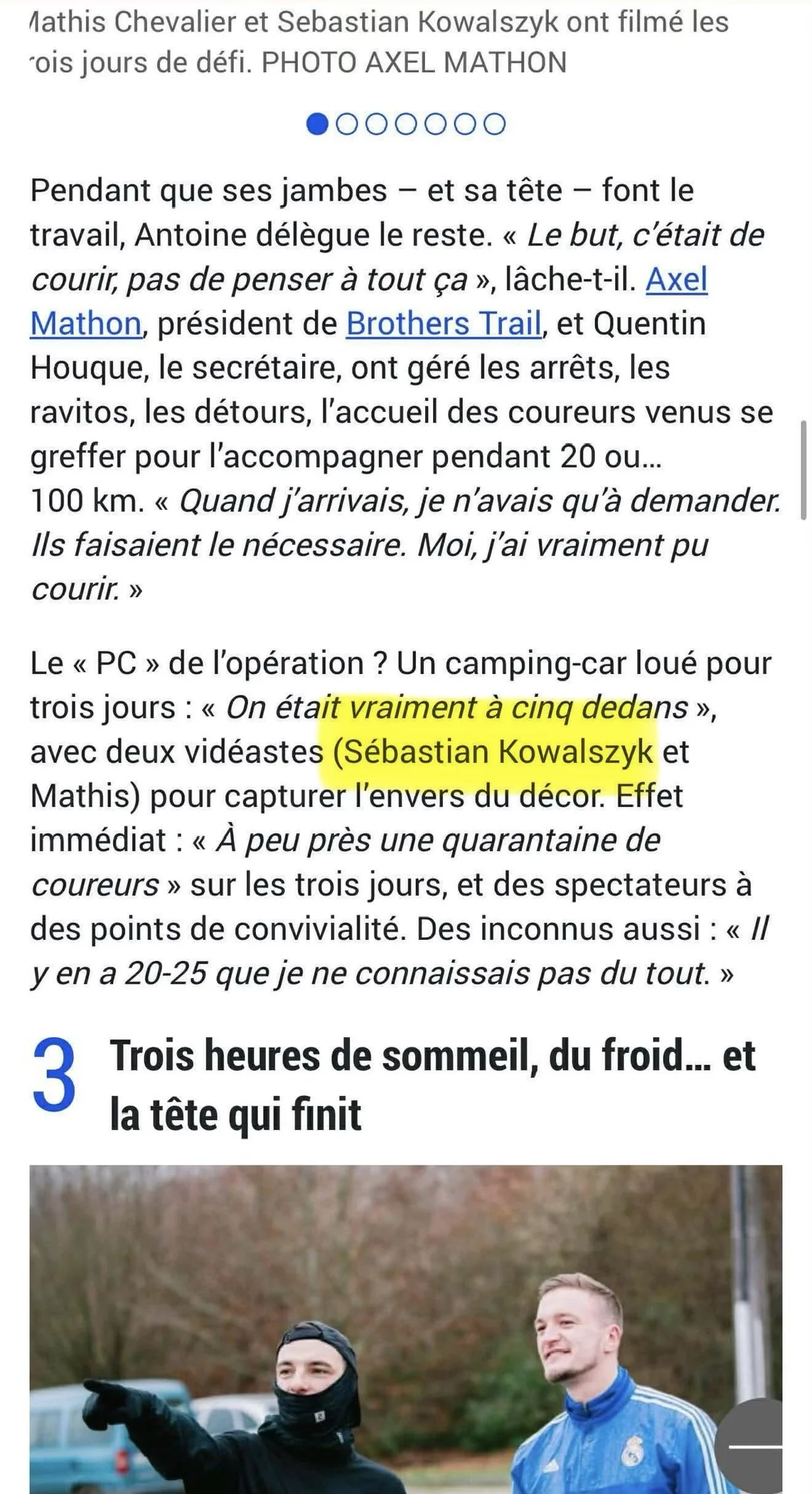 Deux hommes discutent lors d'une séance d'entraînement en extérieur, l'un porte un masque noir et un bonnet, et l'autre porte un maillot de football bleu, avec des arbres en arrière-plan.