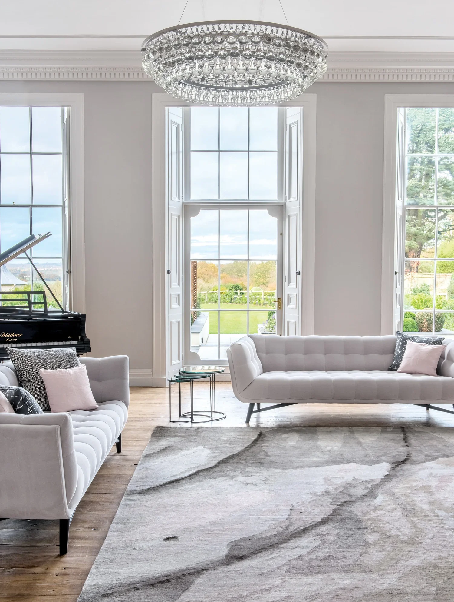 Bright living room with white sofas, a gray patterned rug, large windows with a view of green outdoors, a crystal chandelier, and a black grand piano.