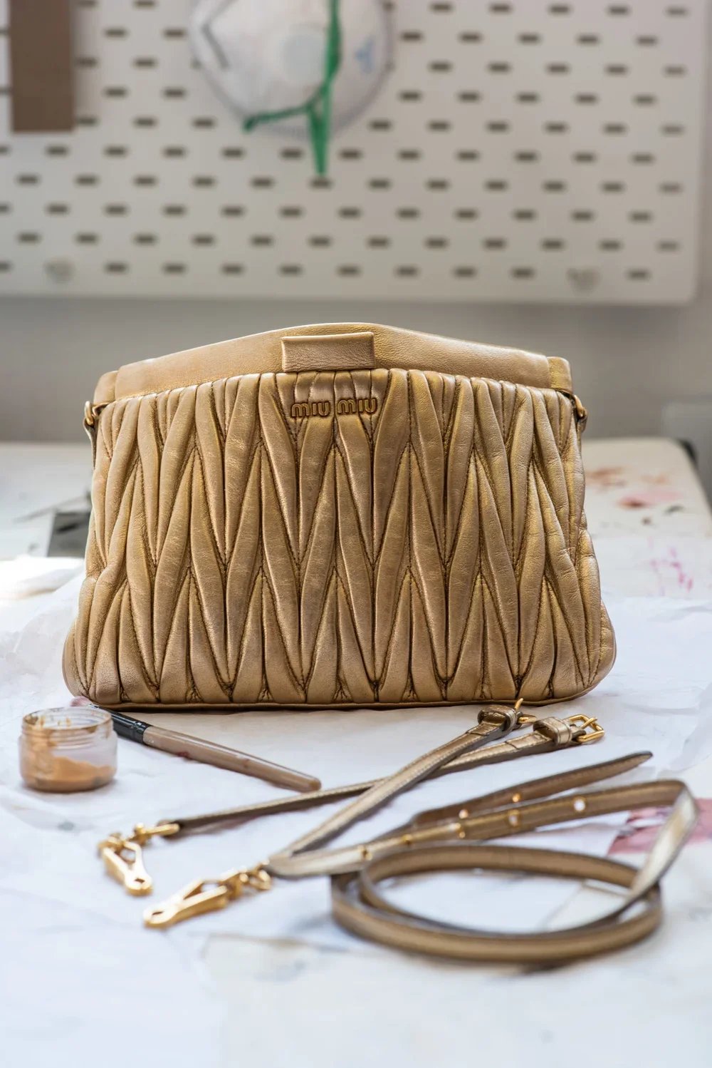 A beige quilted Miu Miu handbag with gold hardware, placed on a white surface with a matching strap and a small container of beige paint, in front of a pegboard with a clock and some papers in the background.