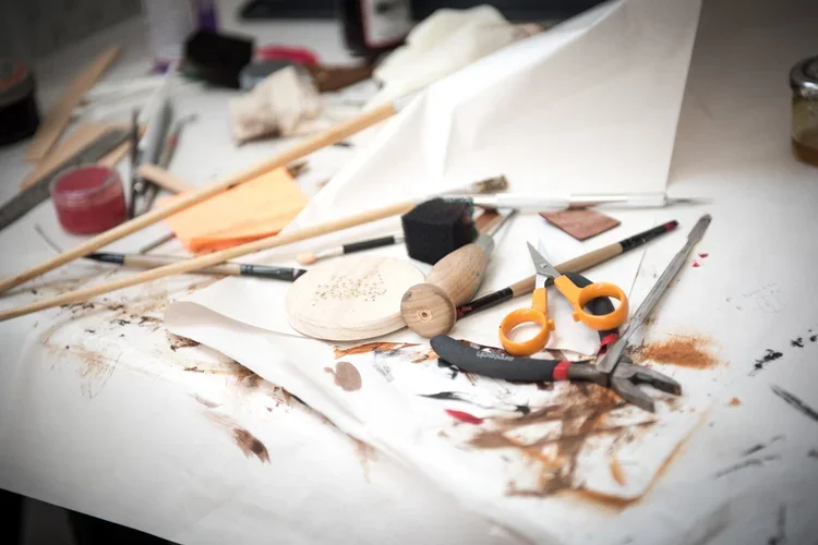 Messy work table with woodworking tools, paint, scissors, and paper scraps.