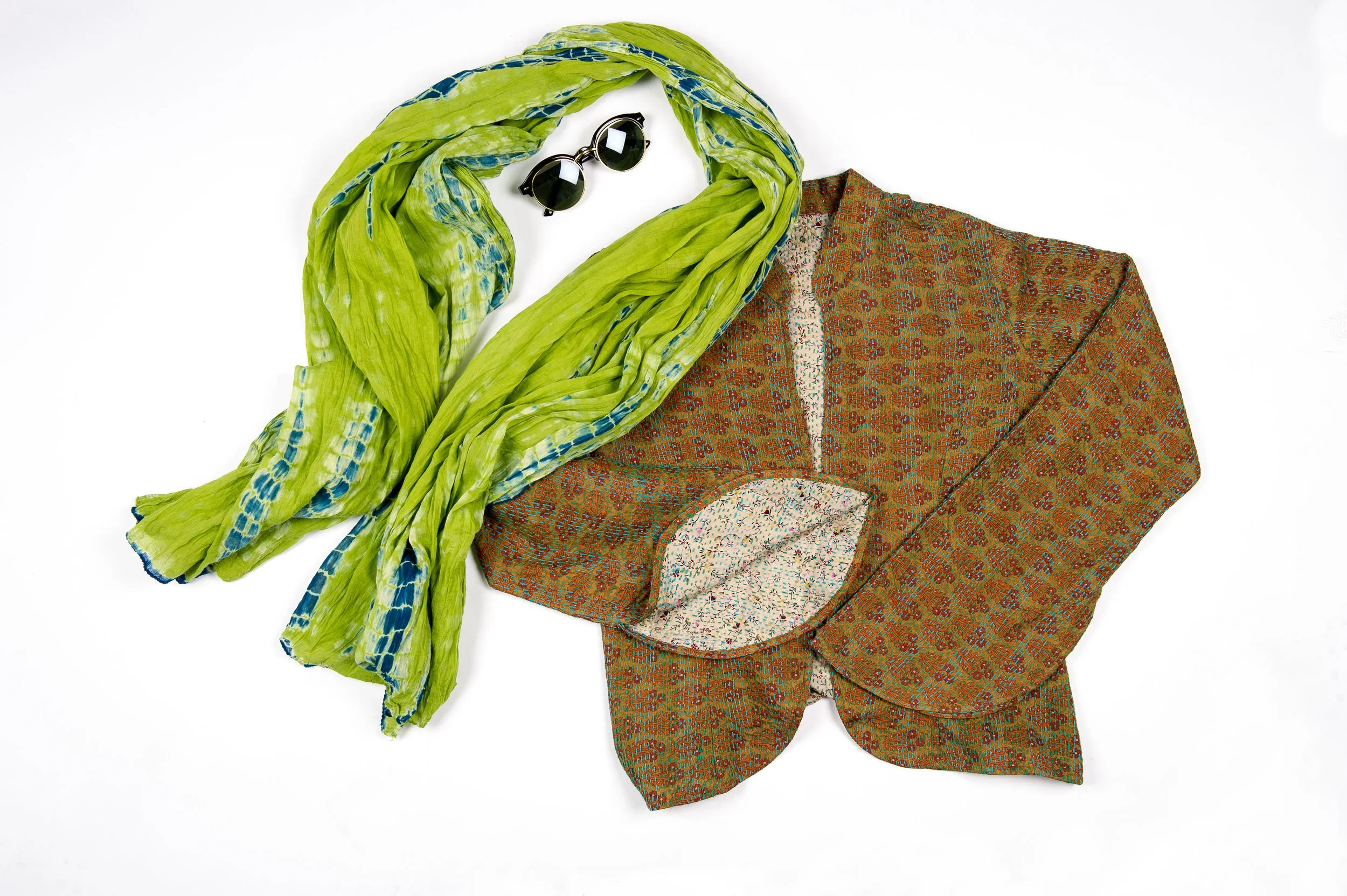 A flat lay of sunglasses, a green scarf with blue accents, and a textured blazer with a floral pattern lining.