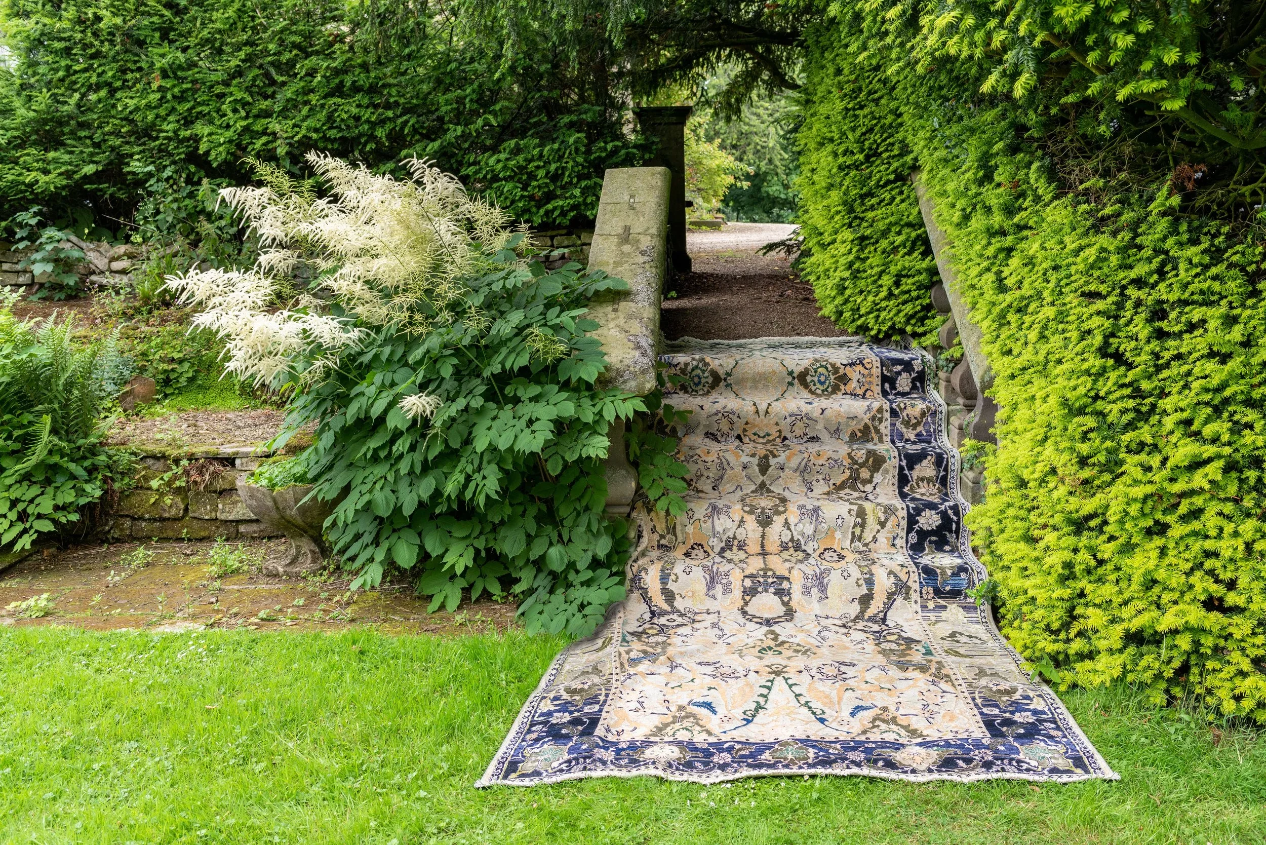 Outdoor scene of a stone staircase covered by a vintage patterned rug, surrounded by lush green plants and bushes.