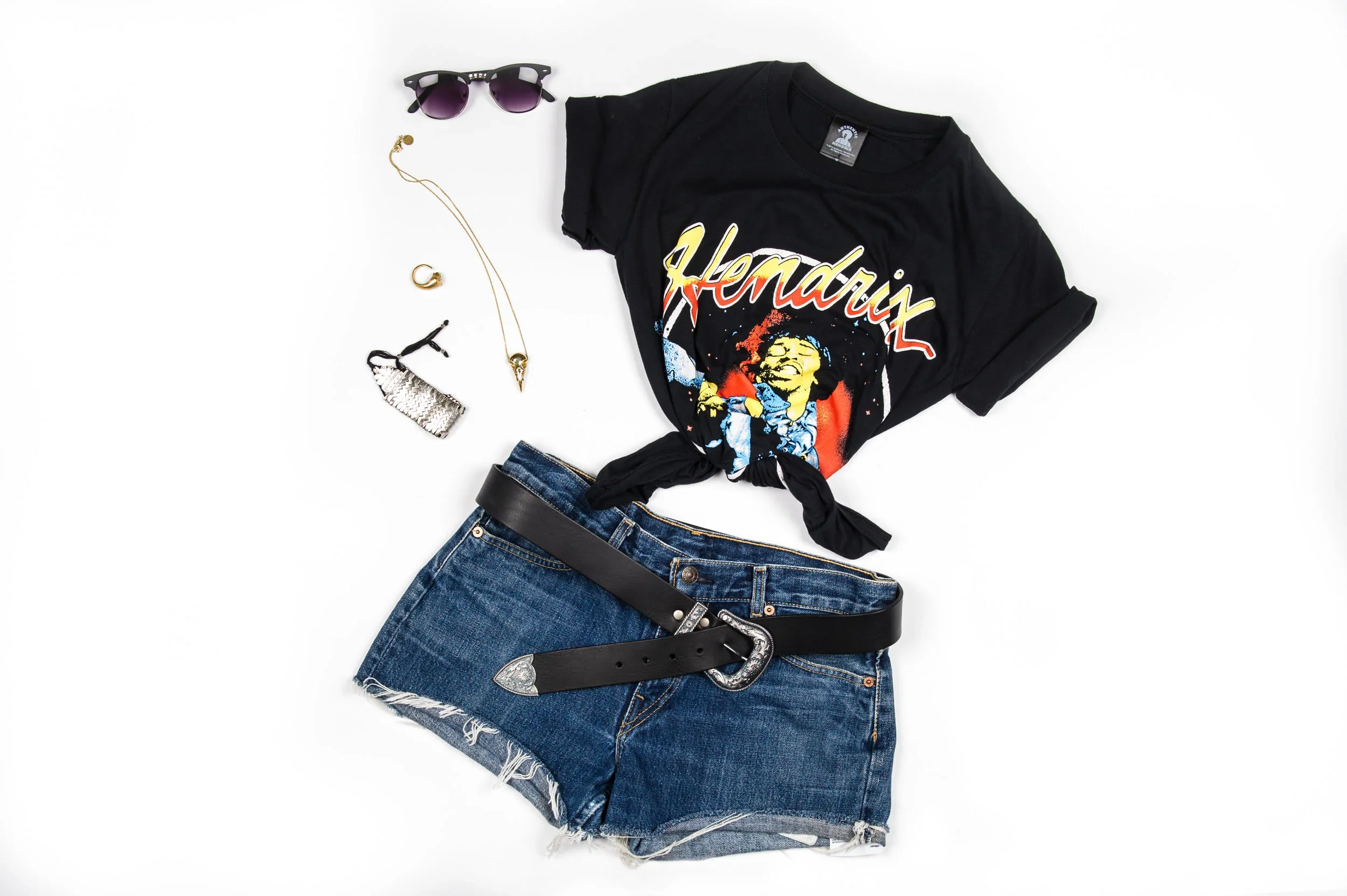 Casual outfit including a black Jimi Hendrix T-shirt tied in front, denim shorts with frayed edges, a black leather belt with a decorative silver buckle, purple sunglasses, a gold necklace with a pendant, a gold ring, a silver ring, and a black hair 