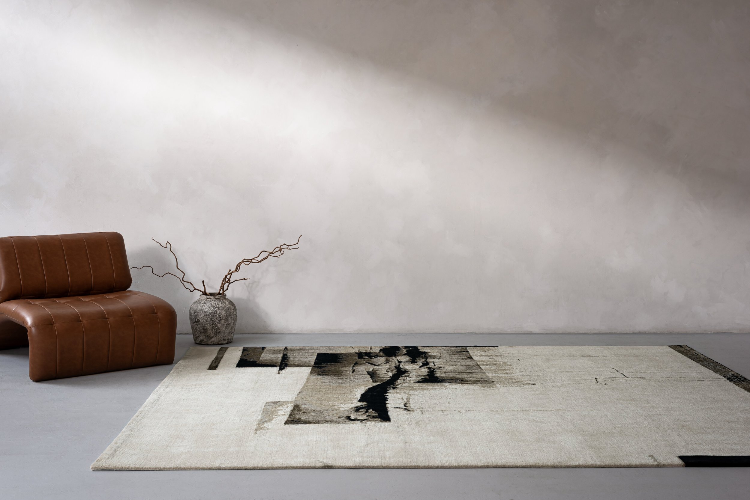 Minimalist living room corner with a brown leather sofa, a textured gray vase with twisted branches, and a modern black, gray, and cream area rug on a light gray floor against a plain light-colored wall.