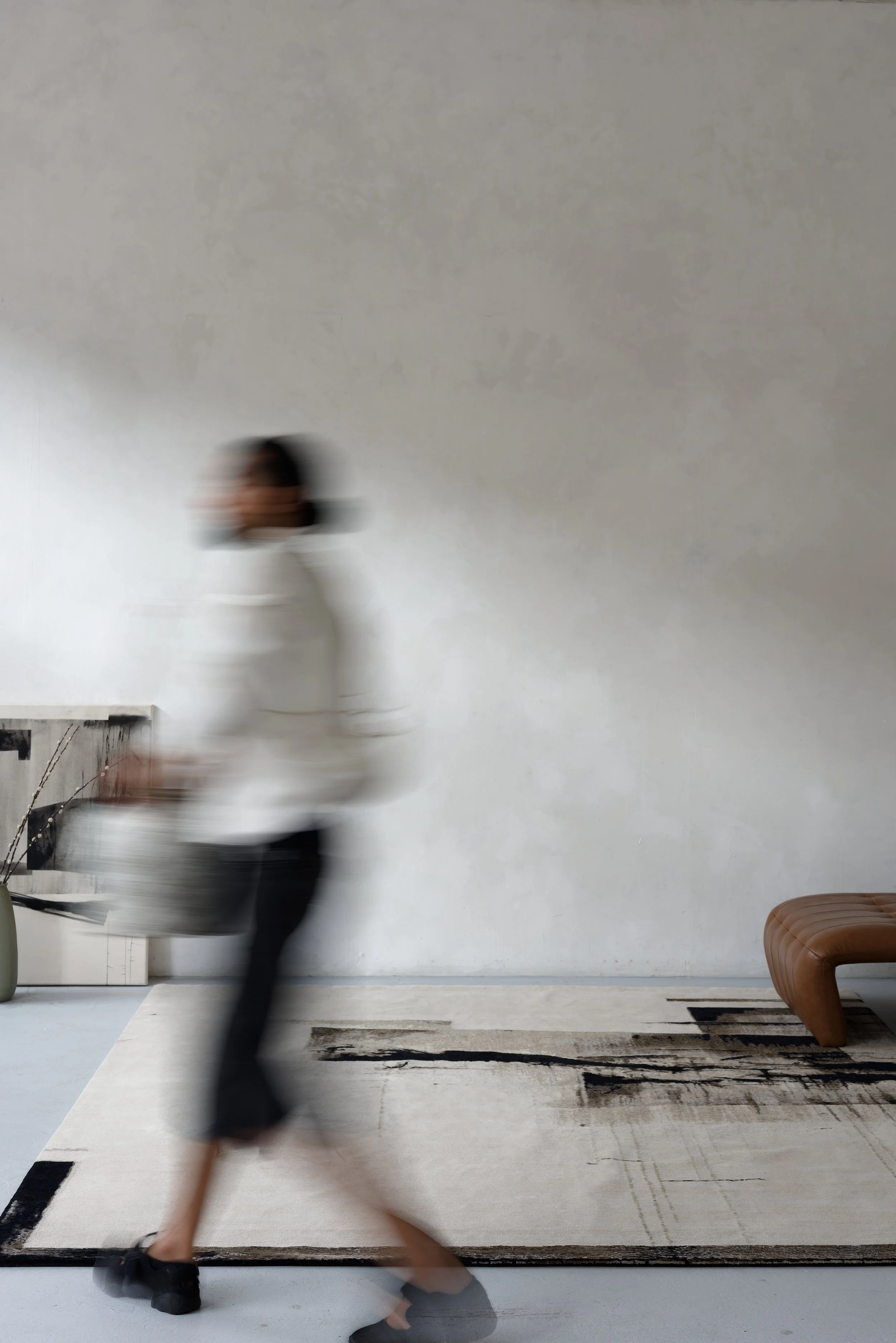 Person walking past a beige and black patterned rug, holding a bag, in a minimalistic room with a white wall and a brown bench.