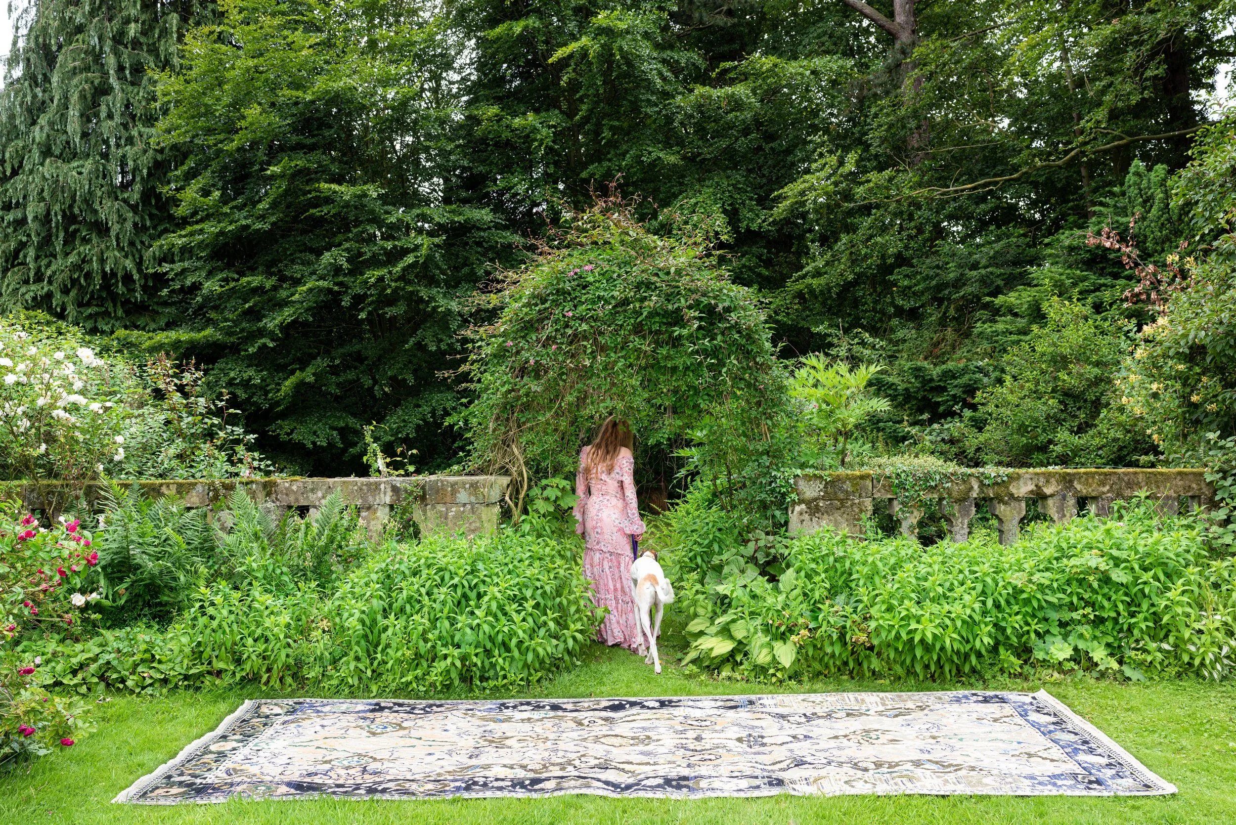 A woman wearing a pink floral dress walking a white dog into a lush green garden with dense trees and bushes, a stone wall, and a decorative outdoor rug on the grass.