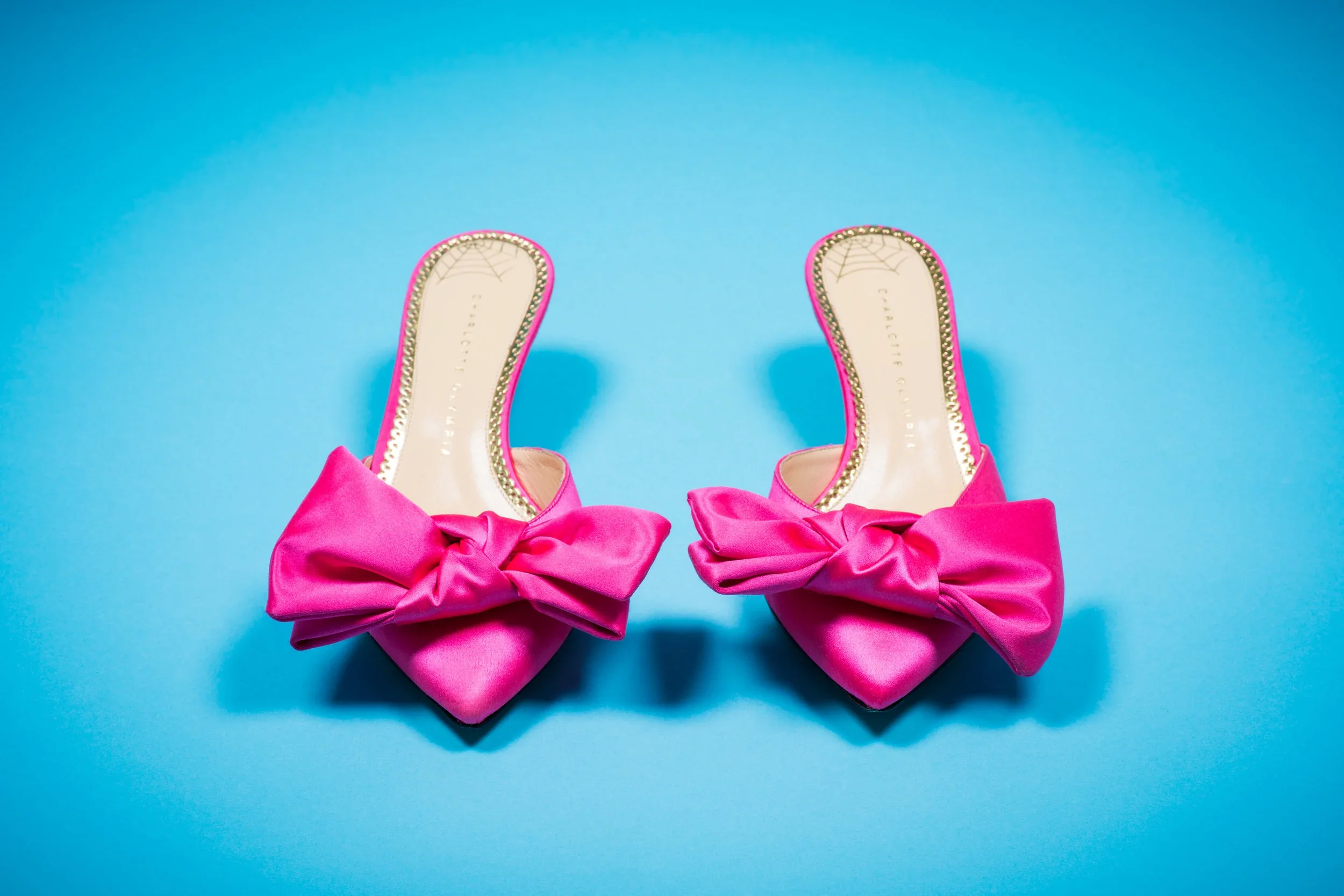 Pair of bright pink high-heeled shoes with large bows on the toes, placed on a solid light blue background.