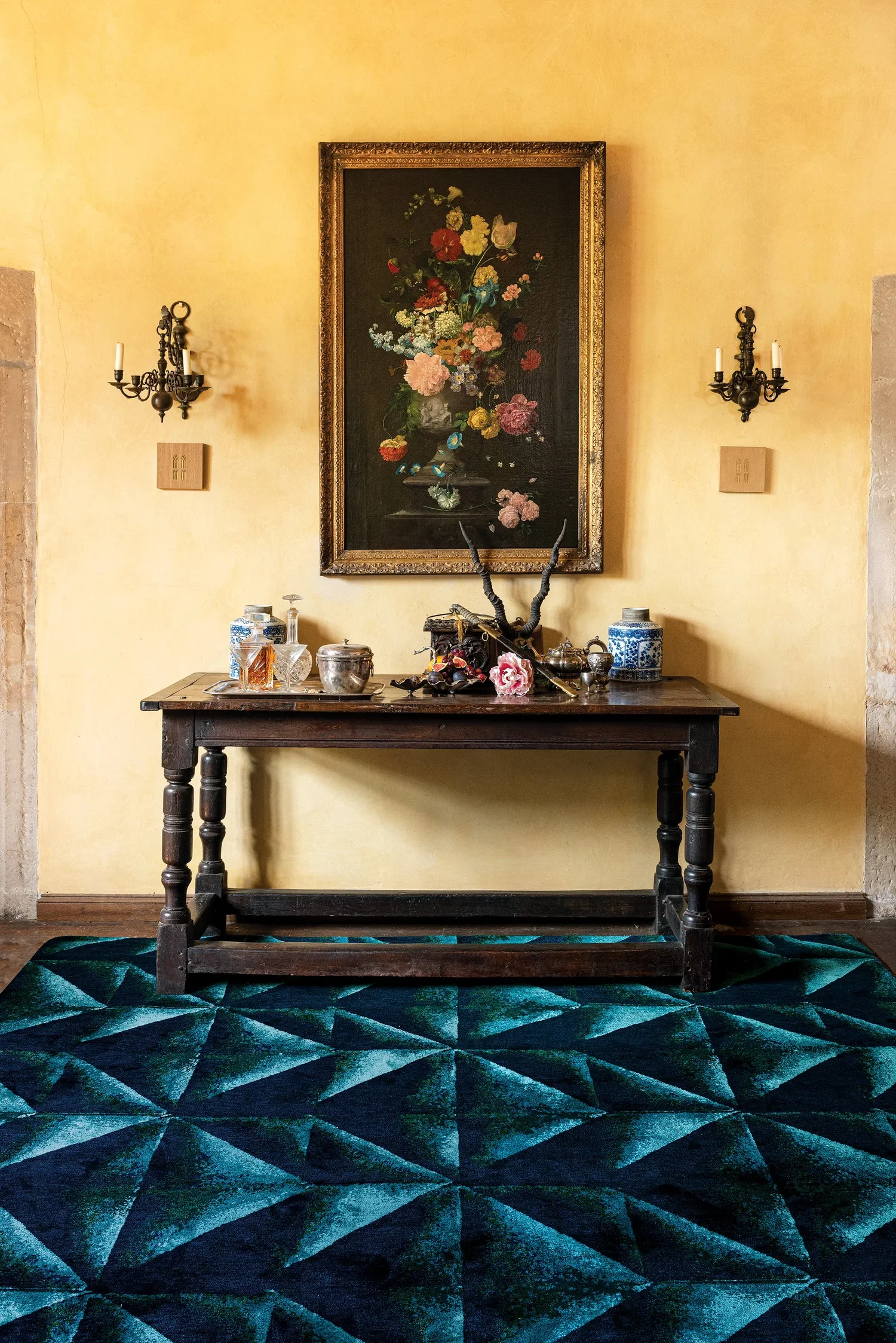 A vintage wooden table against a yellow wall, decorated with various antique containers and objects, with a framed floral still life painting above, and two wall-mounted candle holders.