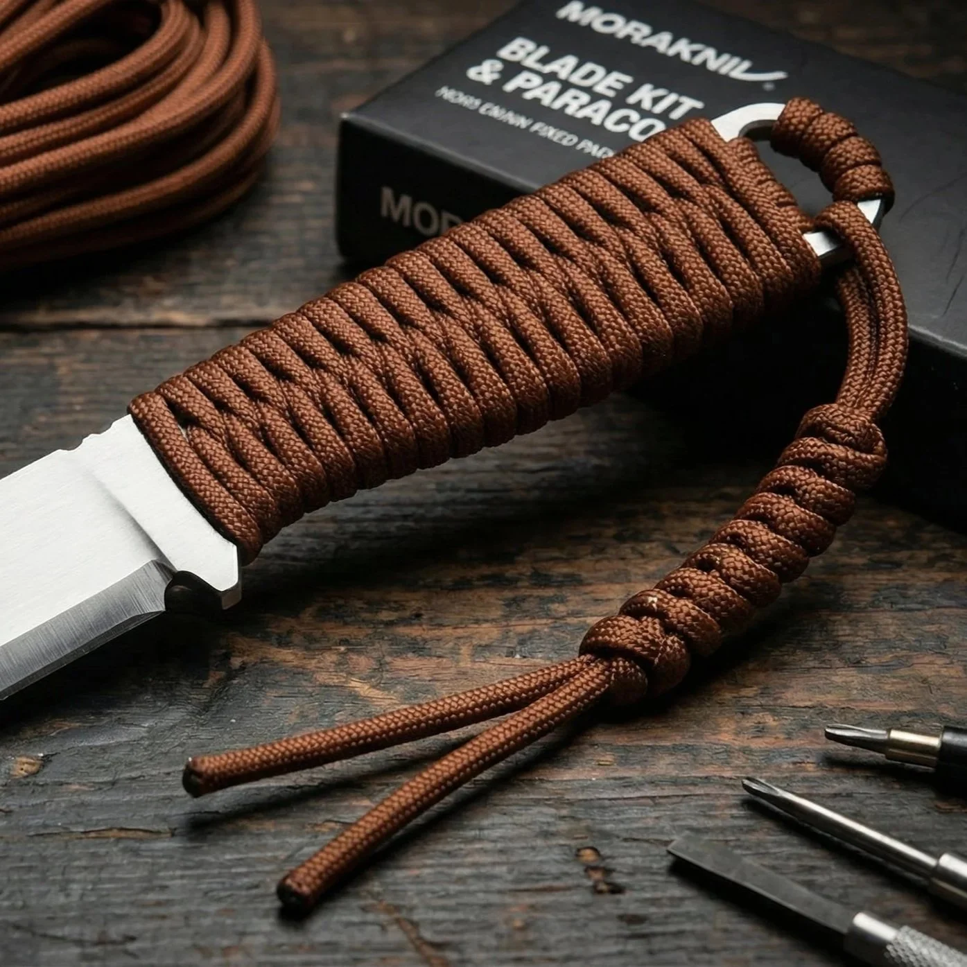 A survival knife with a brown wrapped handle, a lanyard, and metal tools on a wooden surface, with a Morakniv blade kit box in the background.