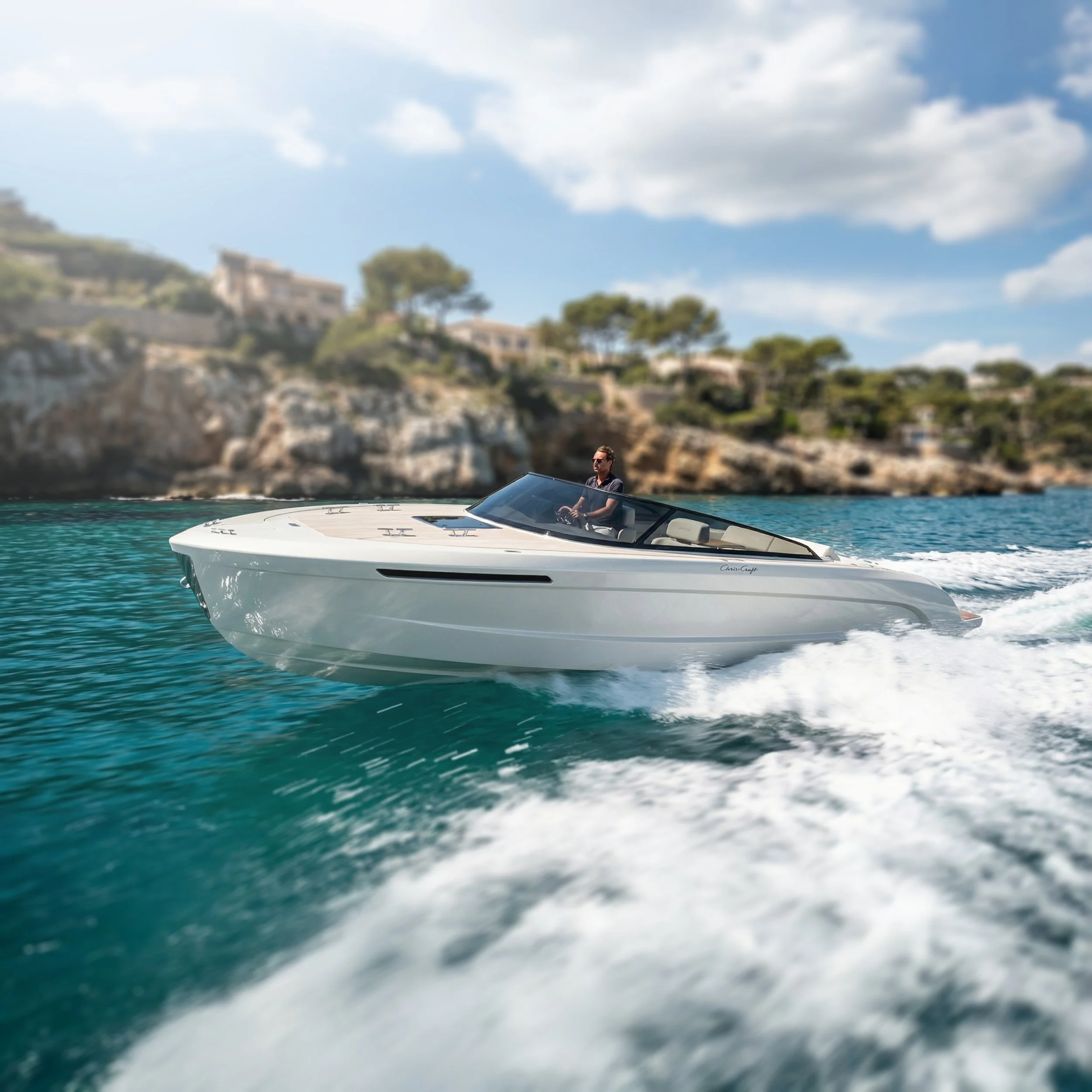 A man sailing a white motorboat on teal waters with a rocky shoreline and trees in the background under a partly cloudy sky.