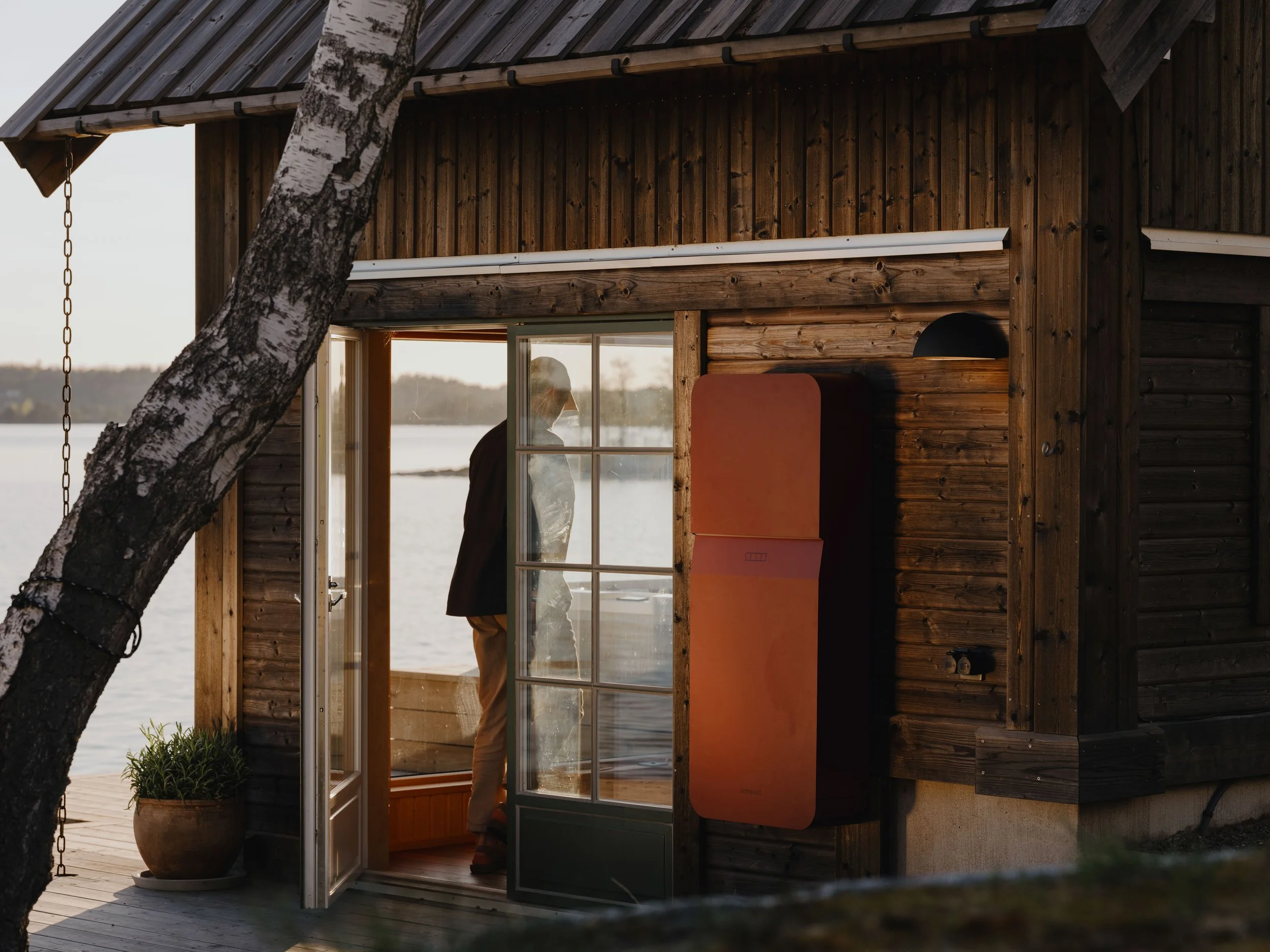 A person stands inside a wooden lakeside cabin looking out through glass-paned door, with the outdoor landscape of water and trees visible.