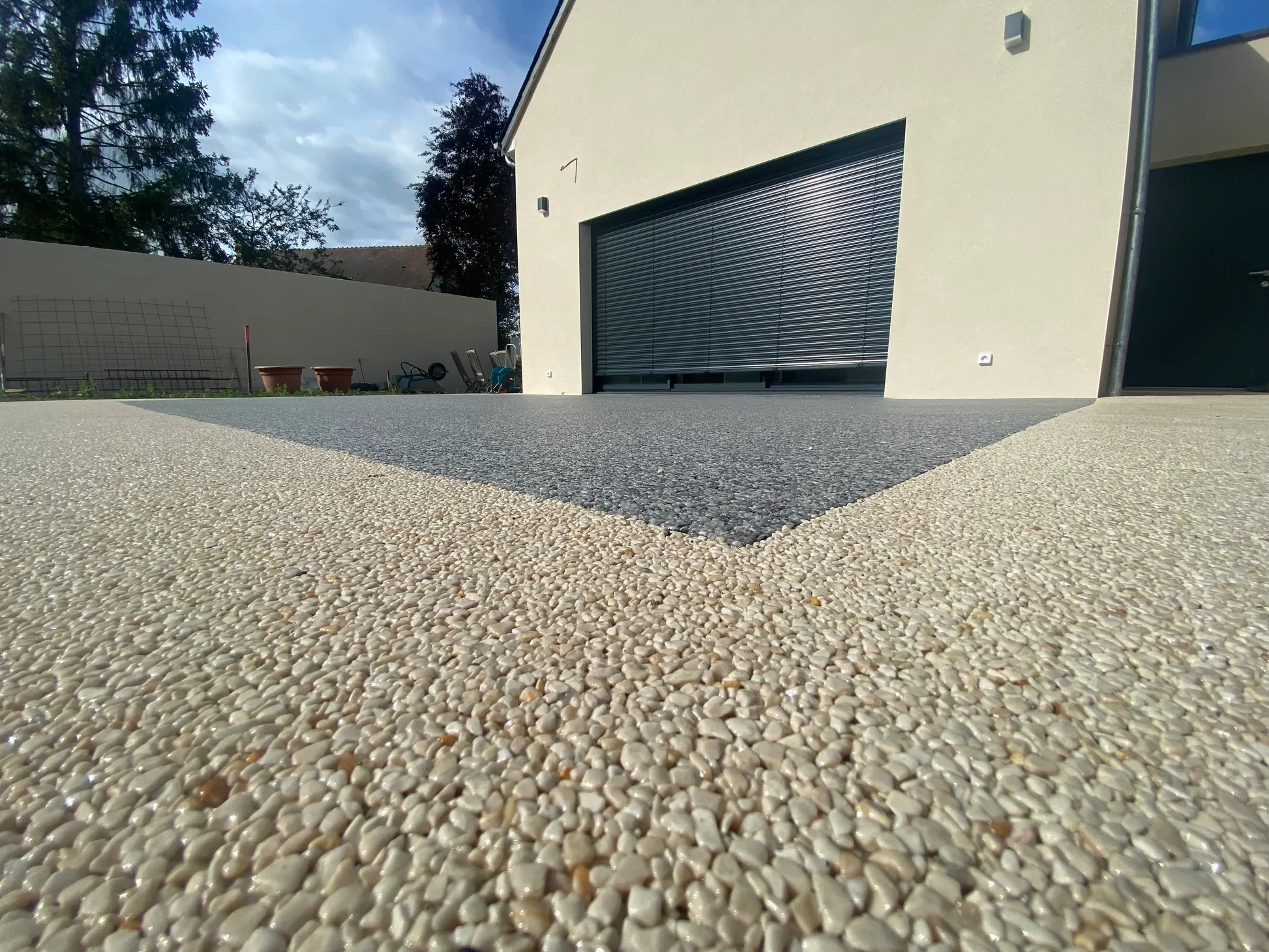 Voûte de pavés en béton à la surface agrégée, en coloris beige clair et gris, devant une maison moderne avec une porte de garage à volet roulant métallique