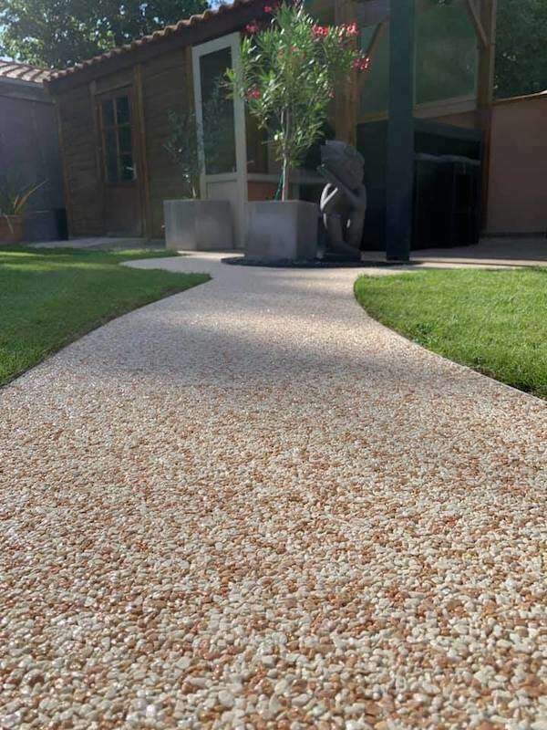 Chemin pavé serpentant dans un jardin devant une petite maison avec une porte en verre et des fenêtres, entouré de gazon vert et d'arbres.