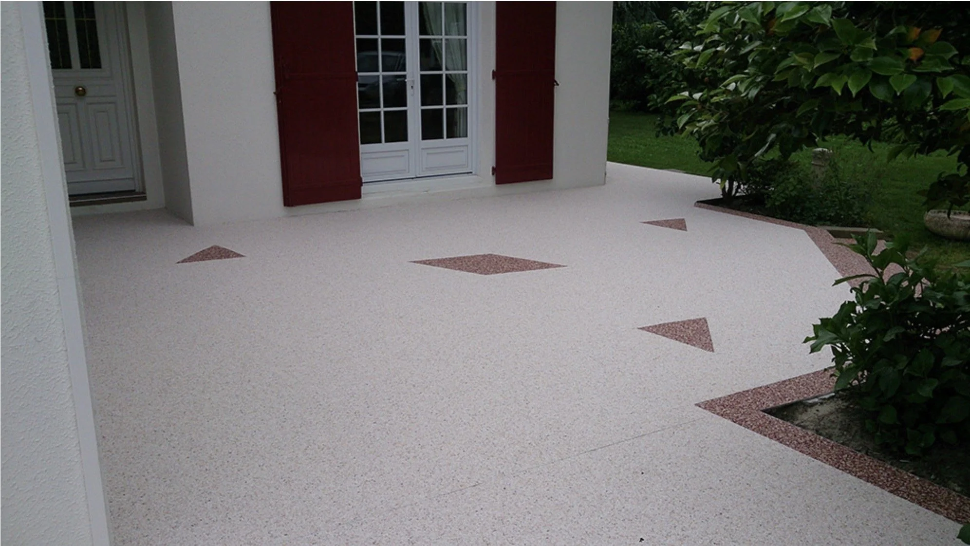 Terrasse devant une maison avec bordures en granit rose, fenêtres avec volets rouges et des plantes vertes environnantes.