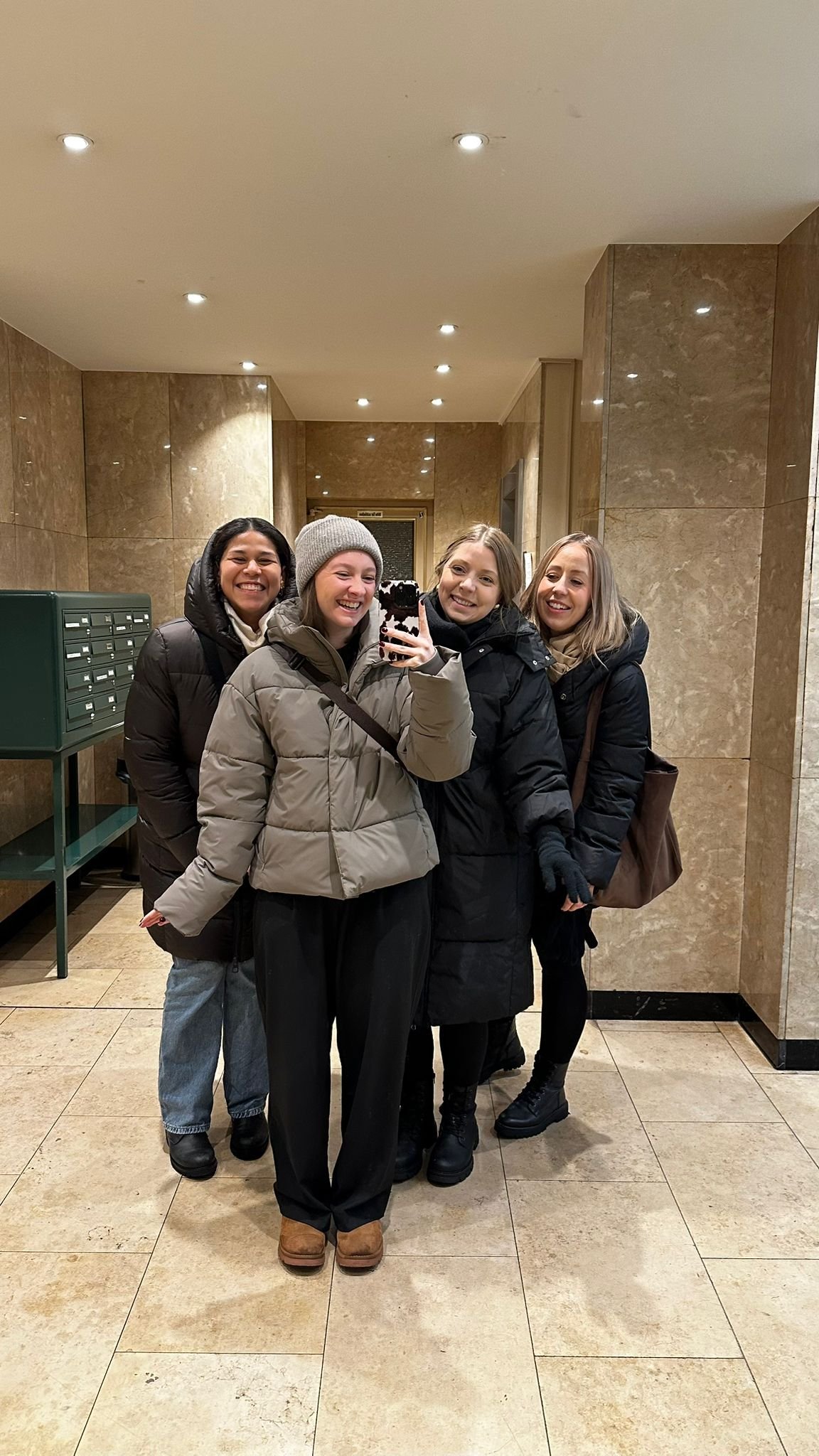 Vier Frauen in Winterkleidung nehmen Selfie in einem Aufzug auf, hinter ihnen sind Briefkästen in einer Lobby.