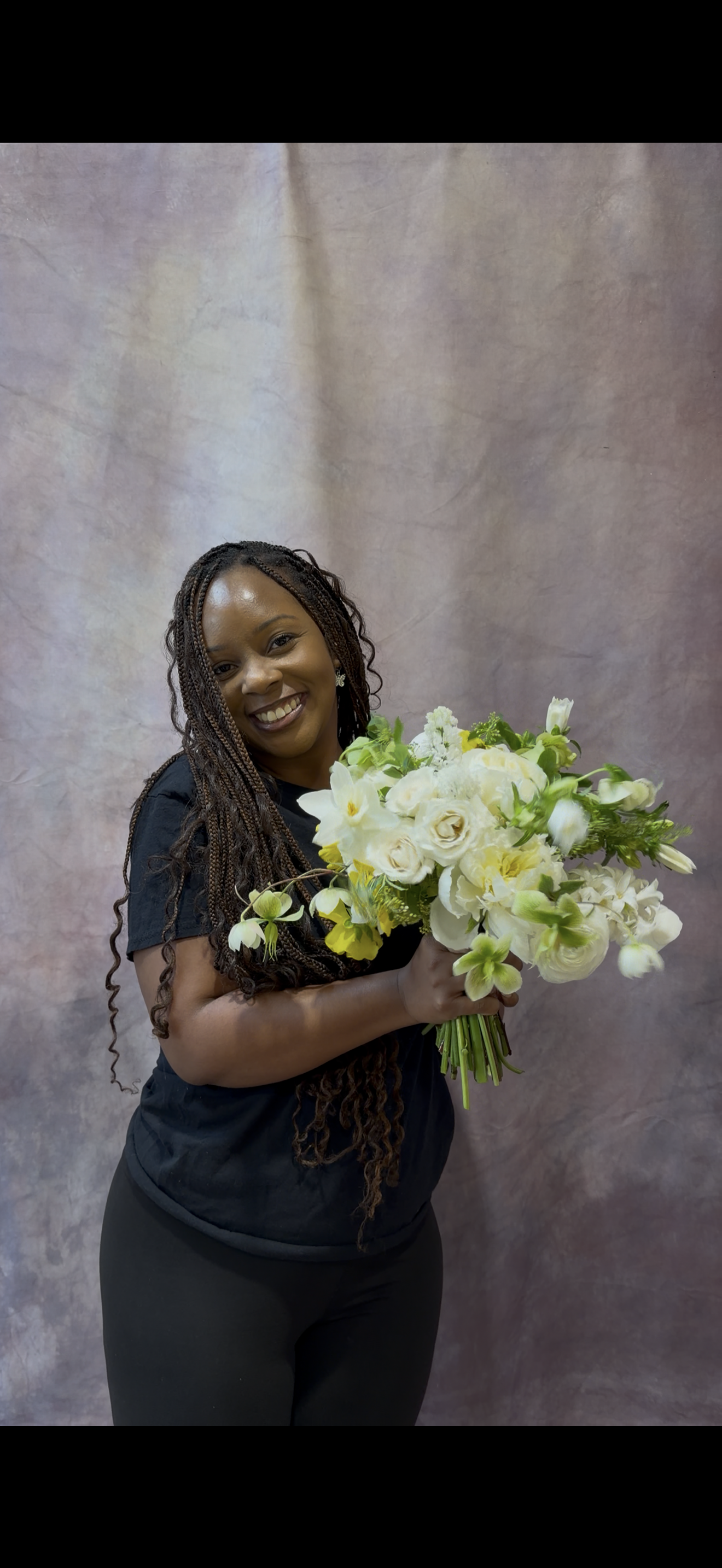 Jay is smiling and holding a large bouquet of white and green flowers, standing in front of a neutral-colored backdrop.