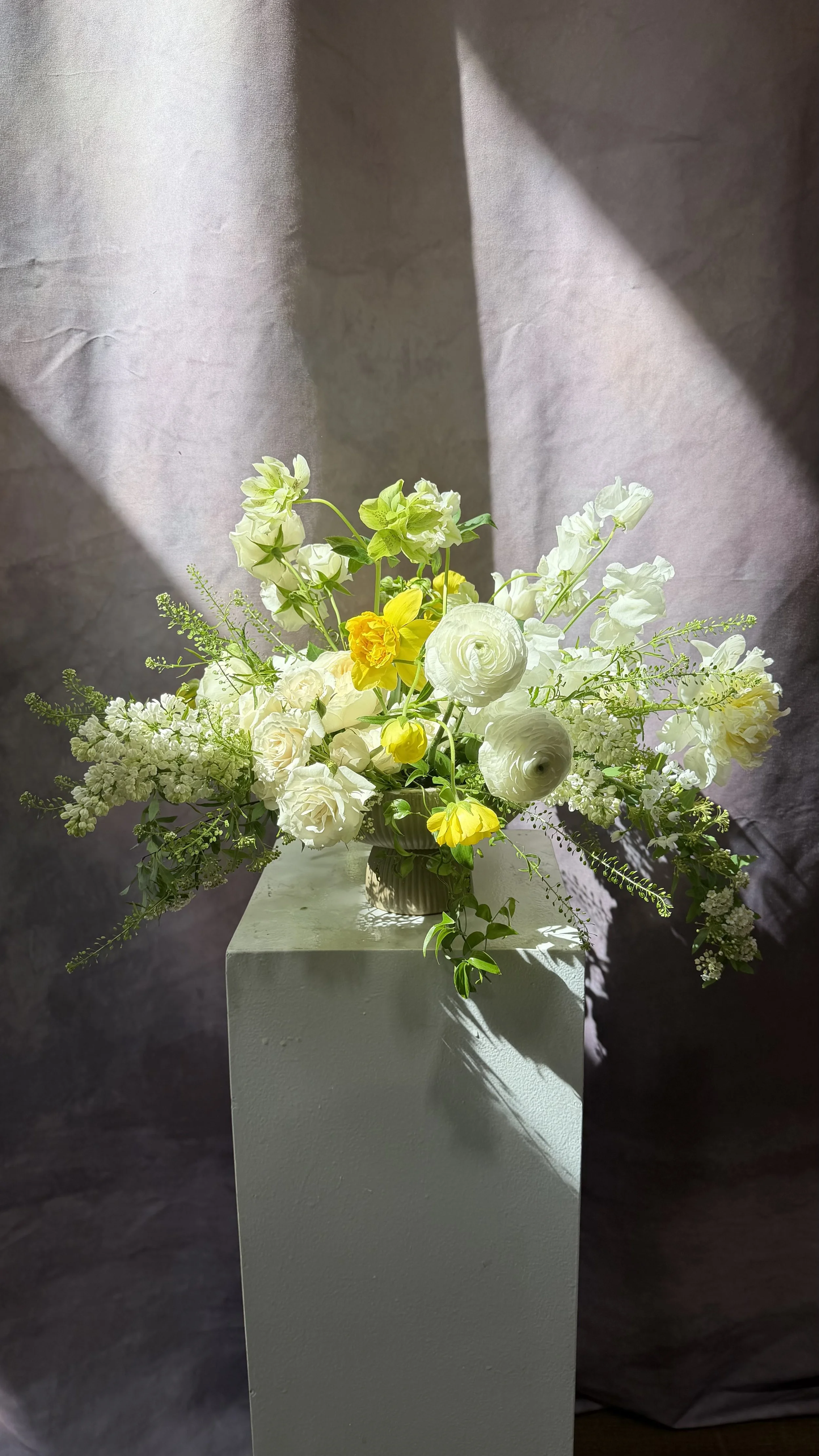 A floral arrangement with white, yellow, and green flowers in a beige vase on a white pedestal.