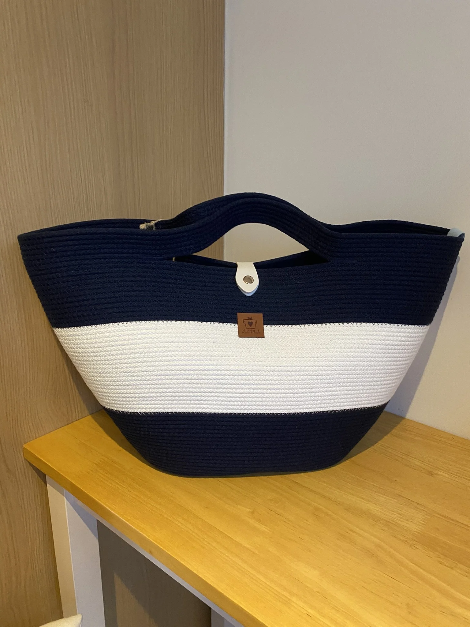 A large tote bag with navy blue, white, and navy horizontal stripes, placed on a wooden surface against a wooden and beige wall.