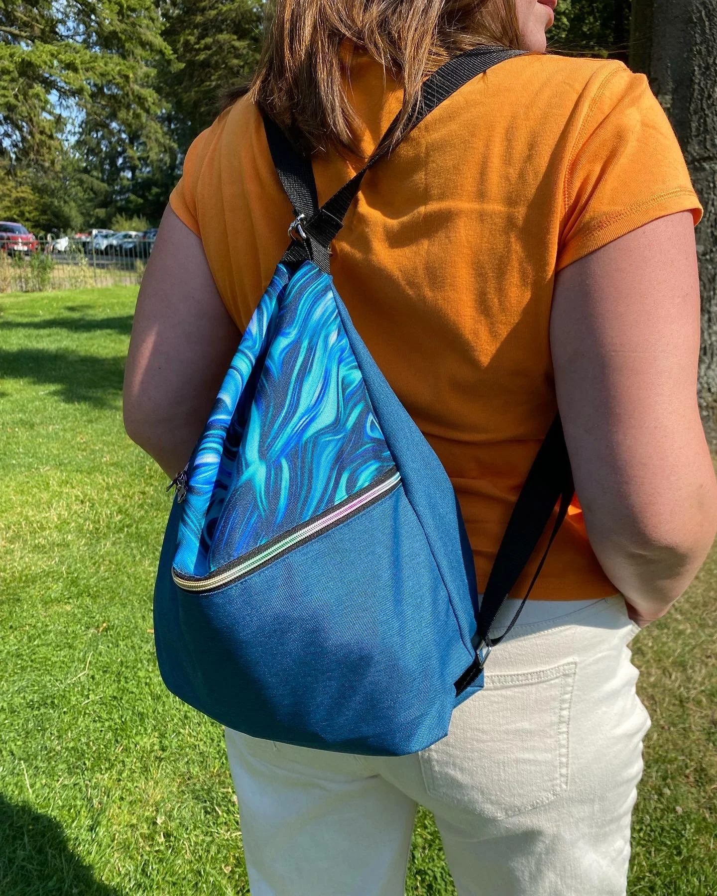 A woman with shoulder-length brown hair wearing an orange T-shirt and white pants, standing outdoors with green grass and trees in the background. She has a blue backpack with a zipper pocket on her back.